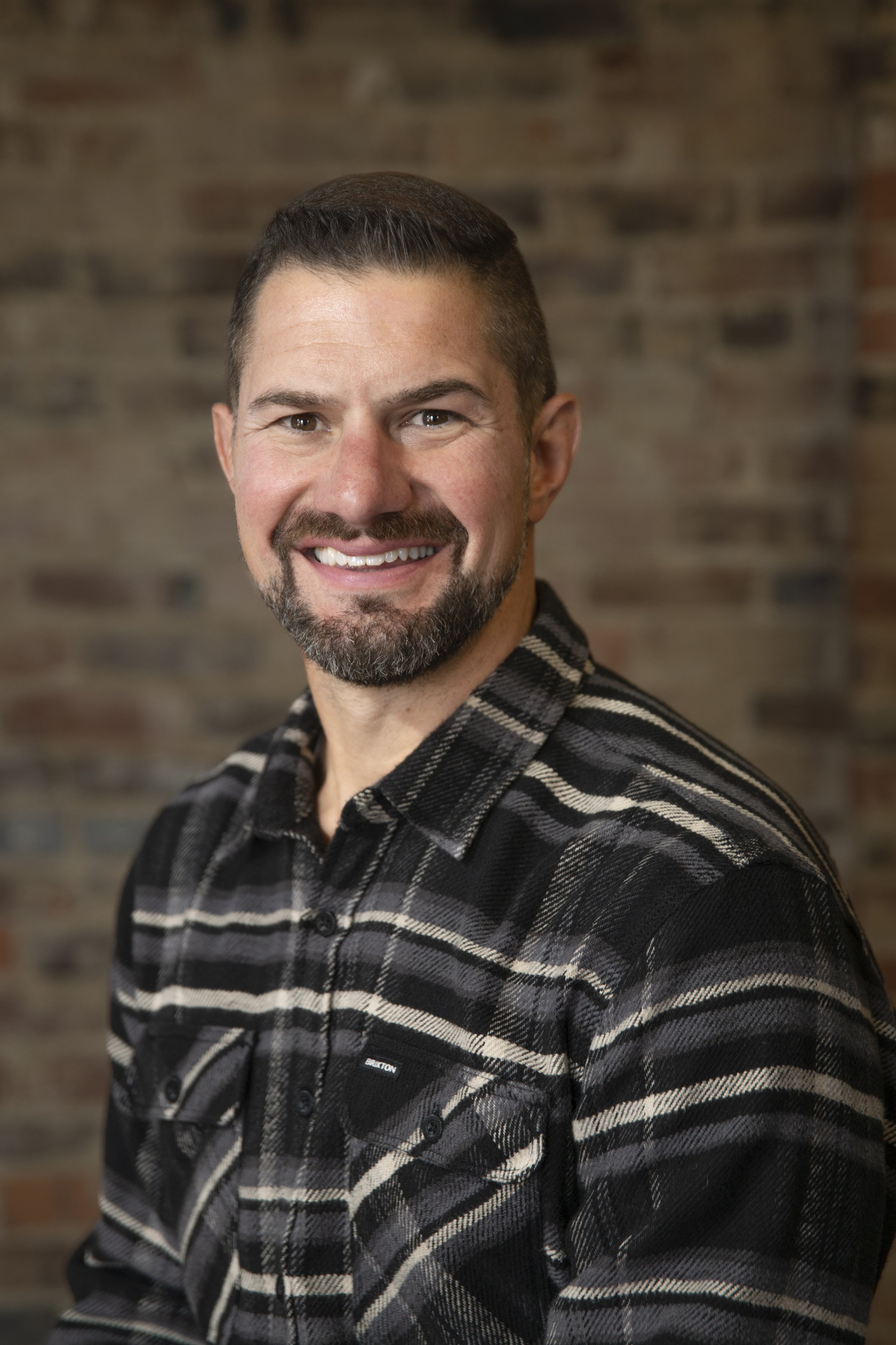 Portrait of a smiling man with short dark hair and a beard, wearing a black and gray plaid shirt, against a brick wall background.