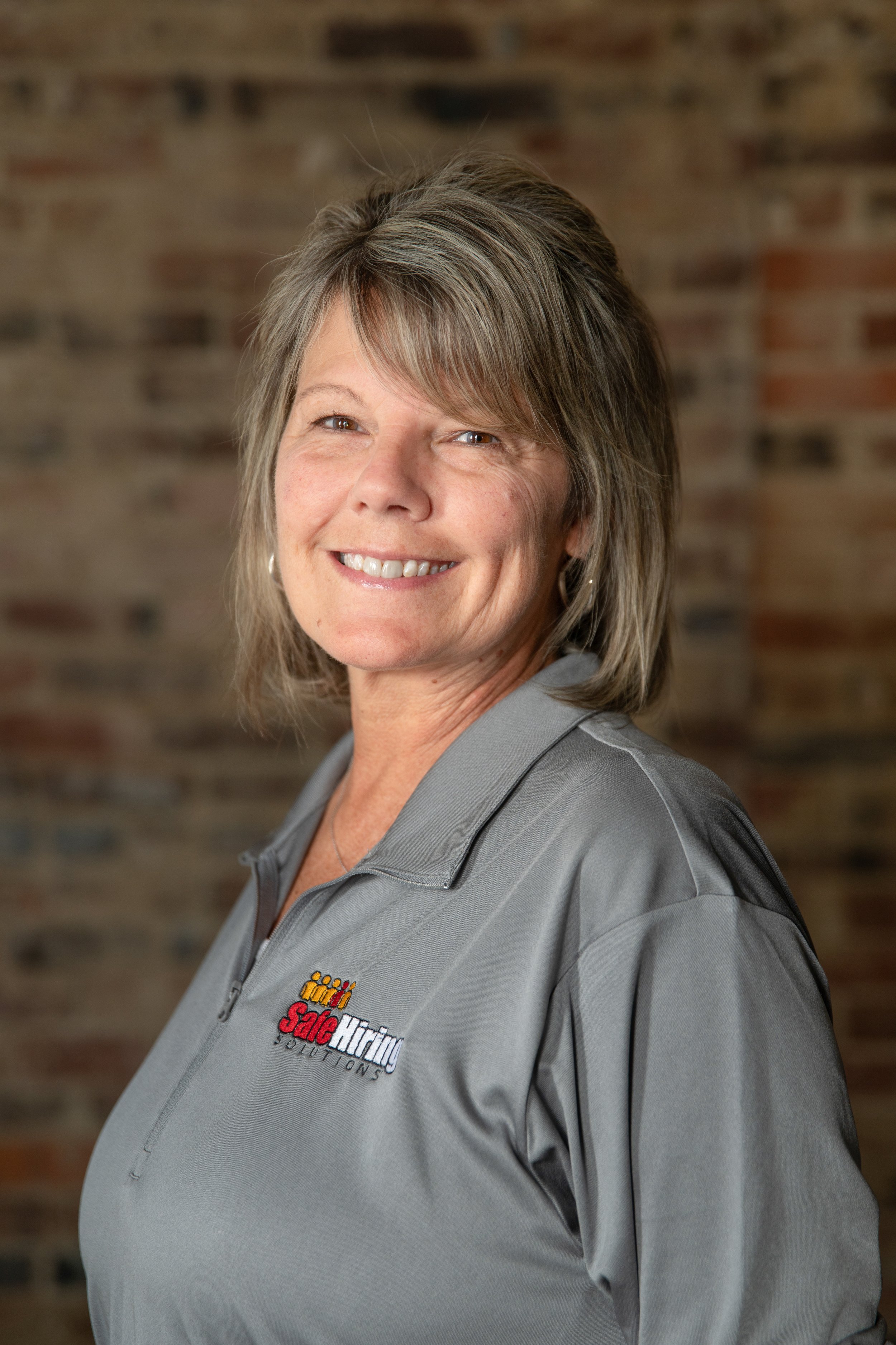 A woman smiling, wearing a gray zipped jacket with a 'Sale Hiring Solutions' logo, standing in front of a brick wall.