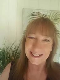 Smiling woman with red hair and bangs, standing indoors near a potted plant, with a framed picture and white wall in the background.