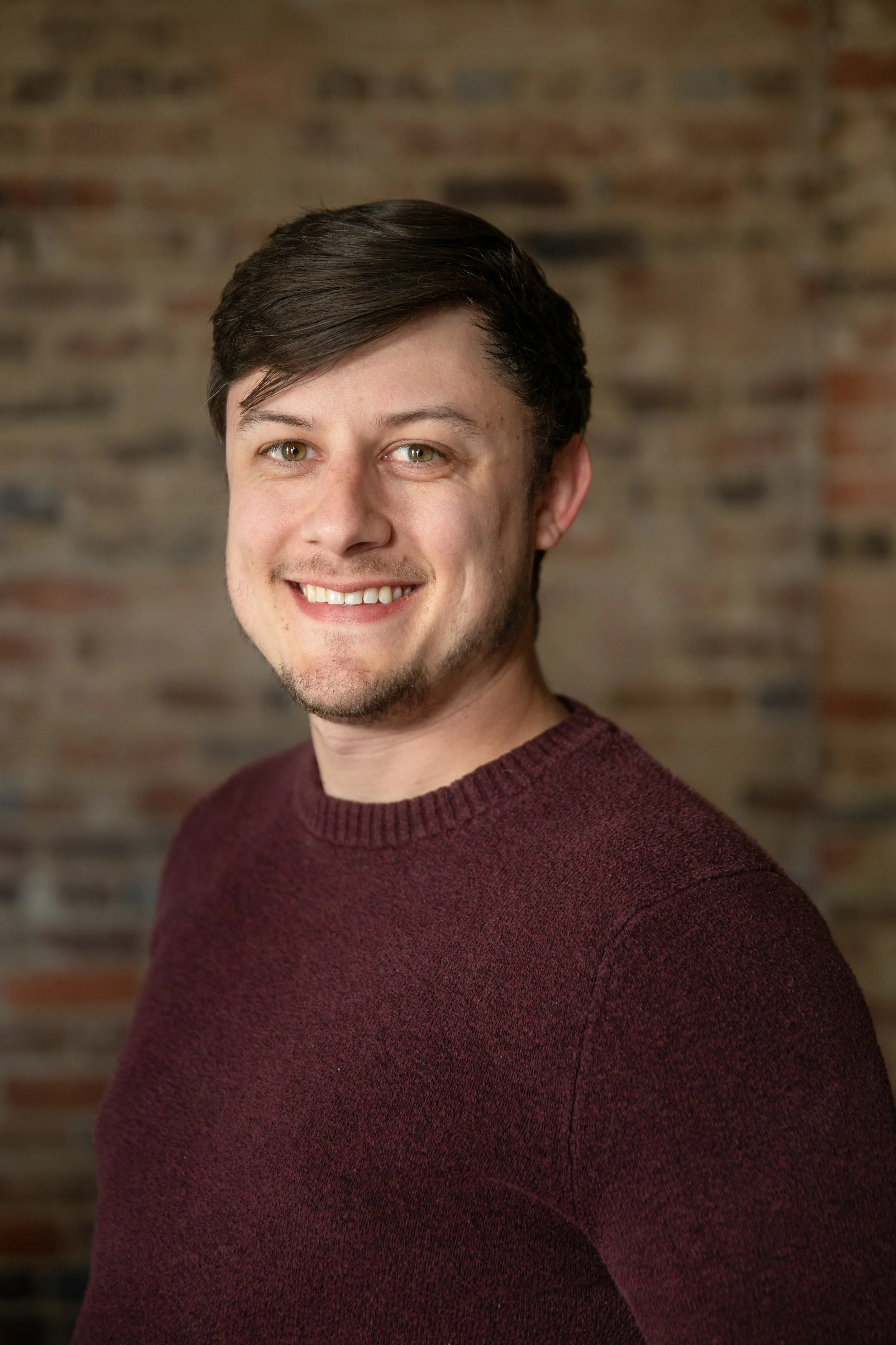 A young man with brown hair, light skin, wearing a maroon sweater, smiling, in front of a blurred brick wall background.