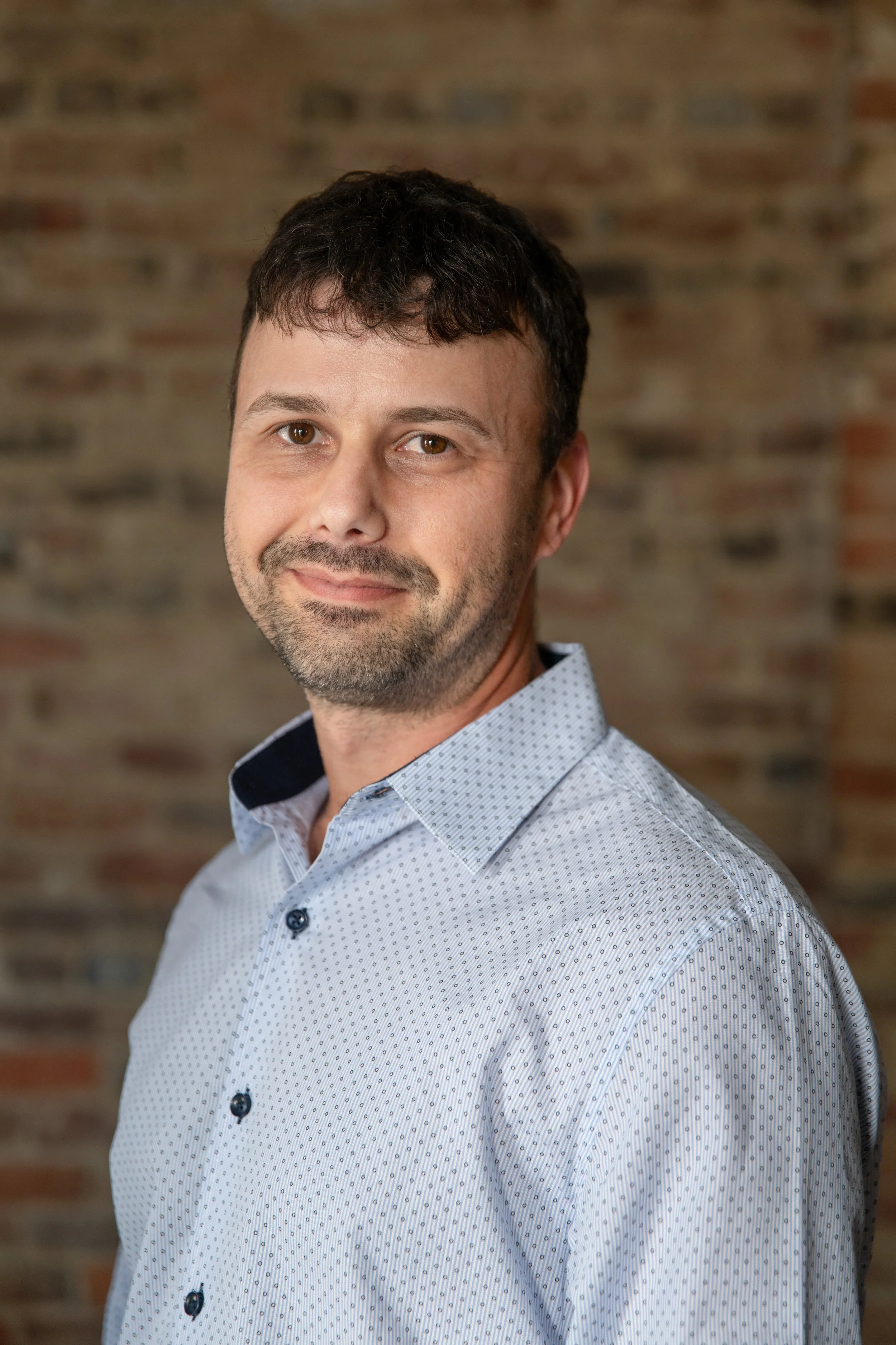 A man with brown hair and a beard wearing a light blue patterned shirt standing in front of a brick wall.