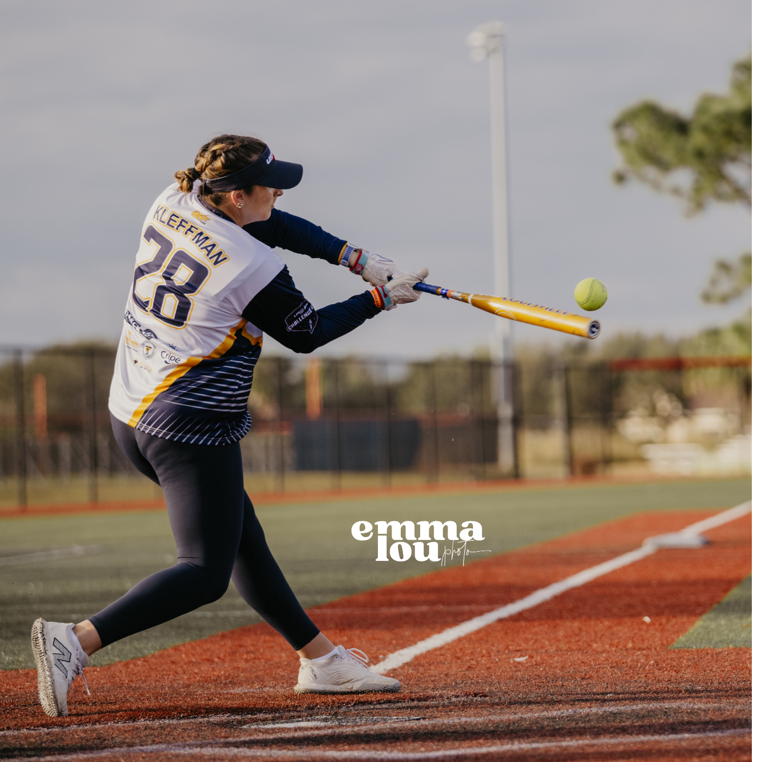 Female softball player in black leggings, white sneakers, and a white and navy uniform with the number 28 and the name KLEFFMAN, swings a yellow bat at a green softball on a field with a fence and trees in the background during daytime.