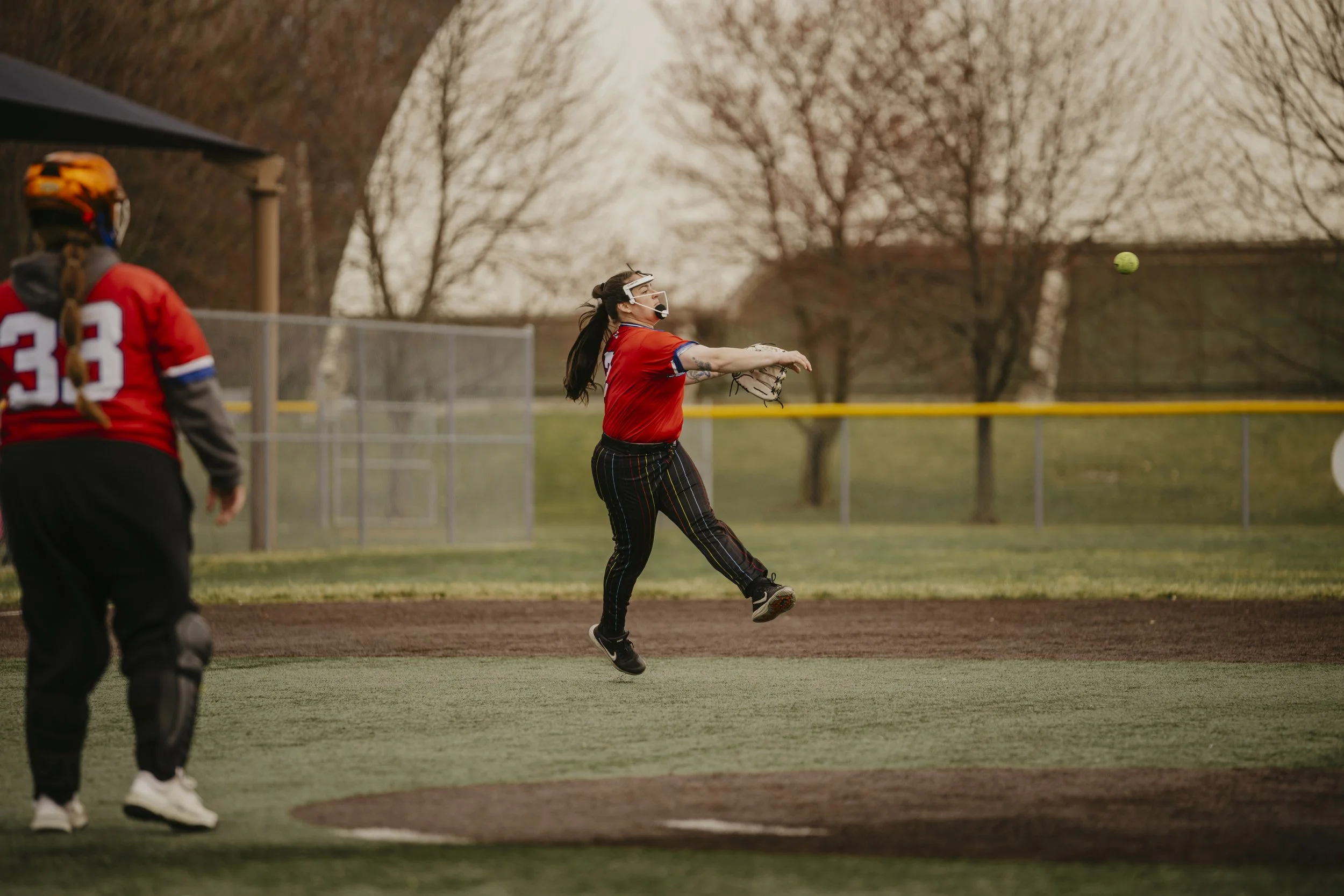 A softball player in red and black gear catches a ball with her glove on a baseball field during daytime, while another player stands nearby, all against a background of trees and yard fencing.