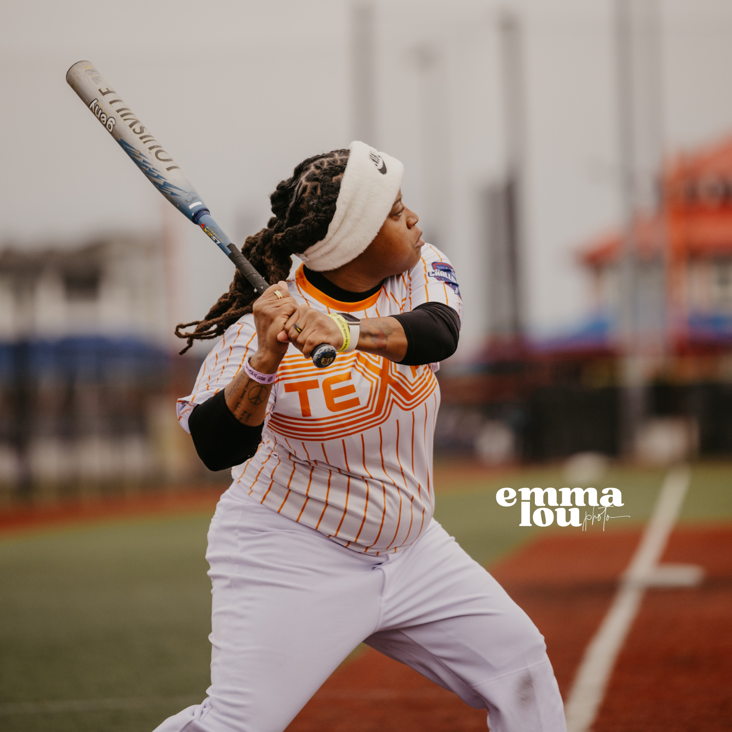 Female softball player in a white and orange uniform, wearing a headband, preparing to swing a bat on the field.
