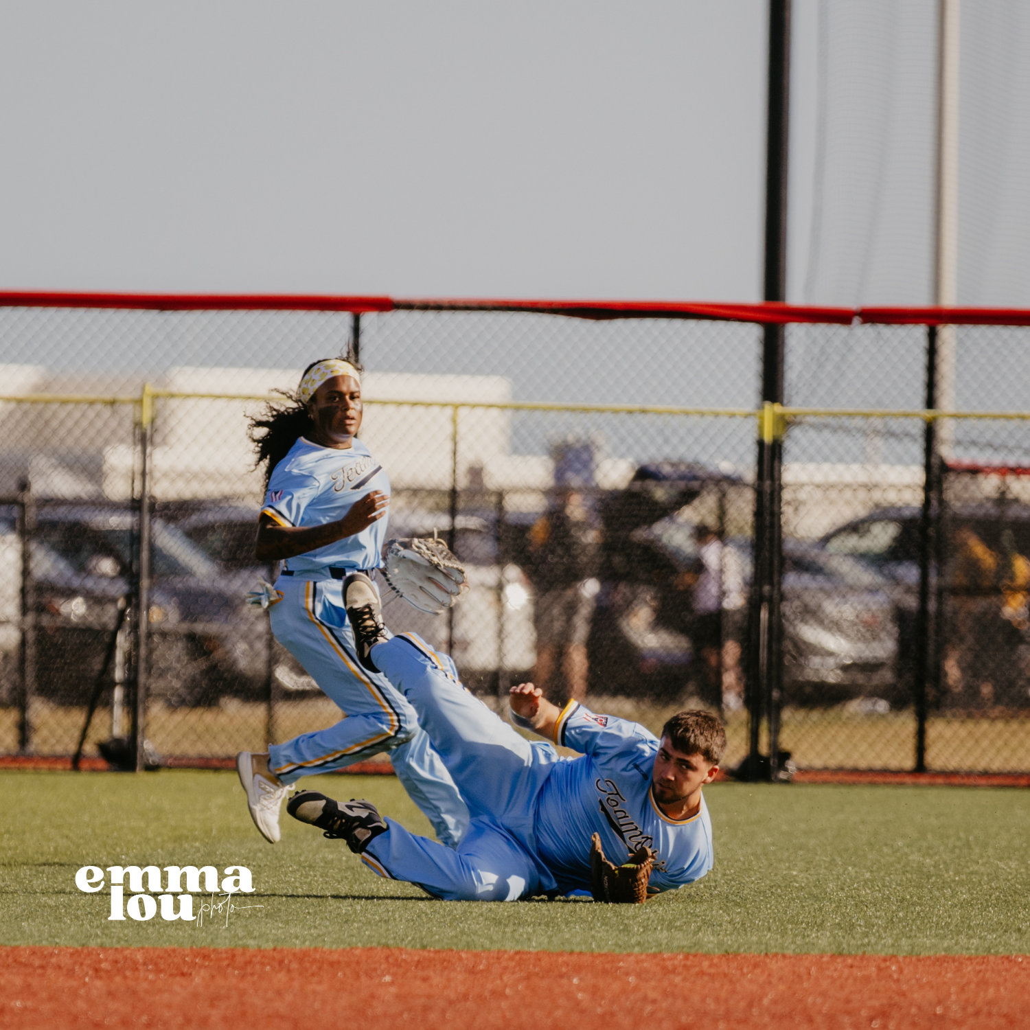 A female softball player in white and blue uniform running past a male player in a similar uniform who is on the ground attempting to catch the ball during a game.