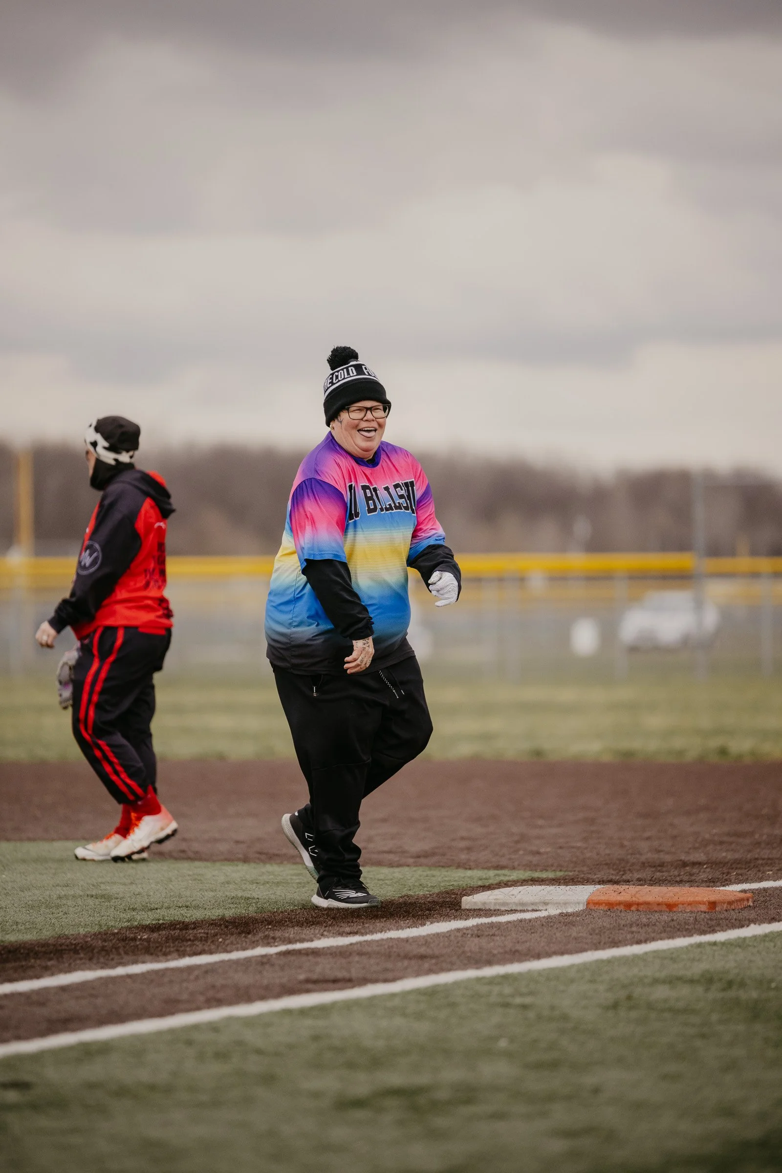 Person smiling and running on a baseball field in cold weather, wearing a colorful sweatshirt, black pants, gloves, and a beanie, with another person in the background.