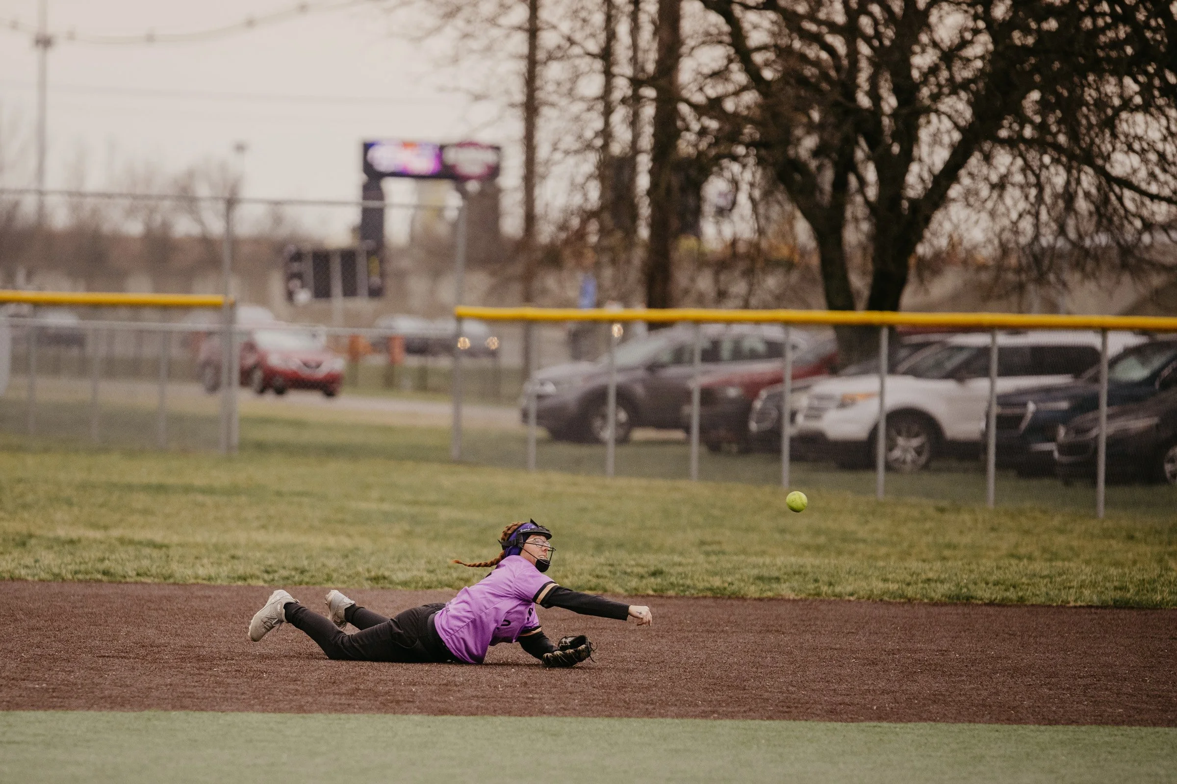 A girl in a purple jersey and black pants diving on the baseball field trying to catch a yellow softball.