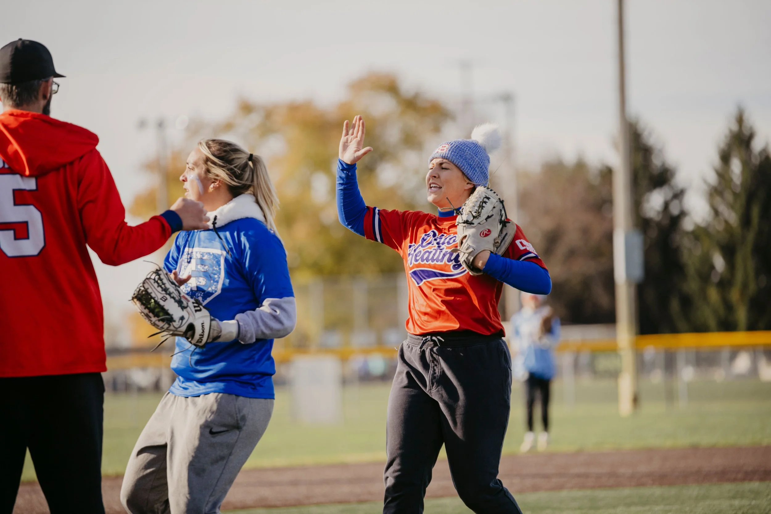 Women softball players on the field, one in red and two in blue, with one woman giving a high five during a game or practice.