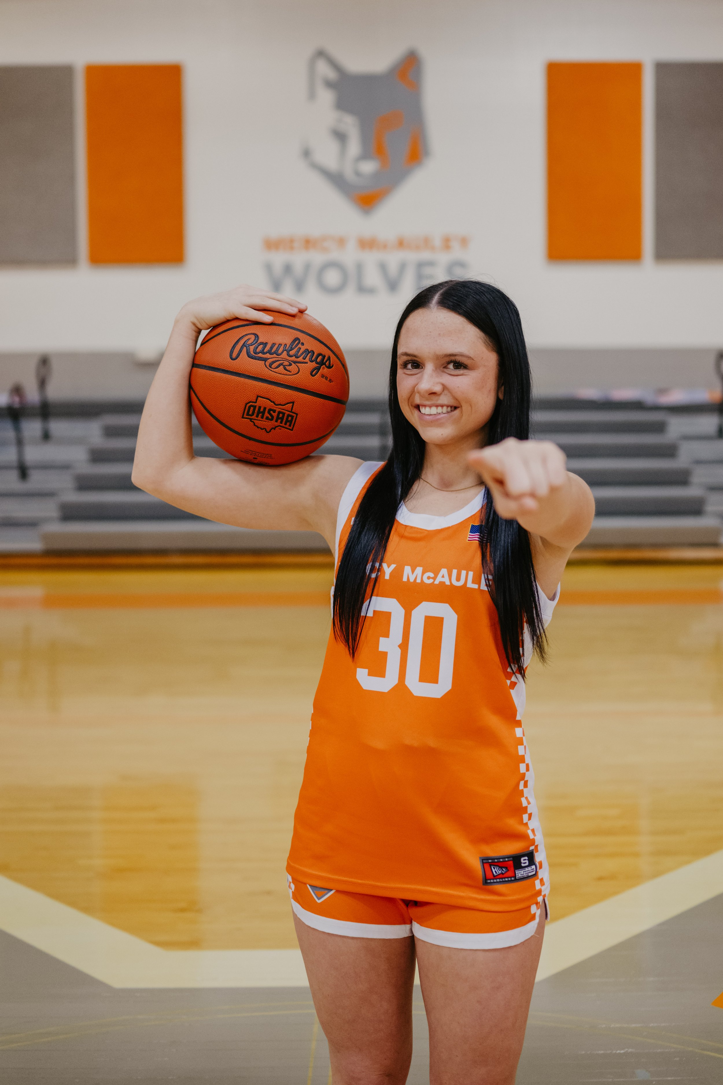 Mercy McAuley Wolves basketball player in orange jersey holding a Rawlings basketball on her shoulder while pointing at the camera with the Wolves logo visible on the gym wall behind her