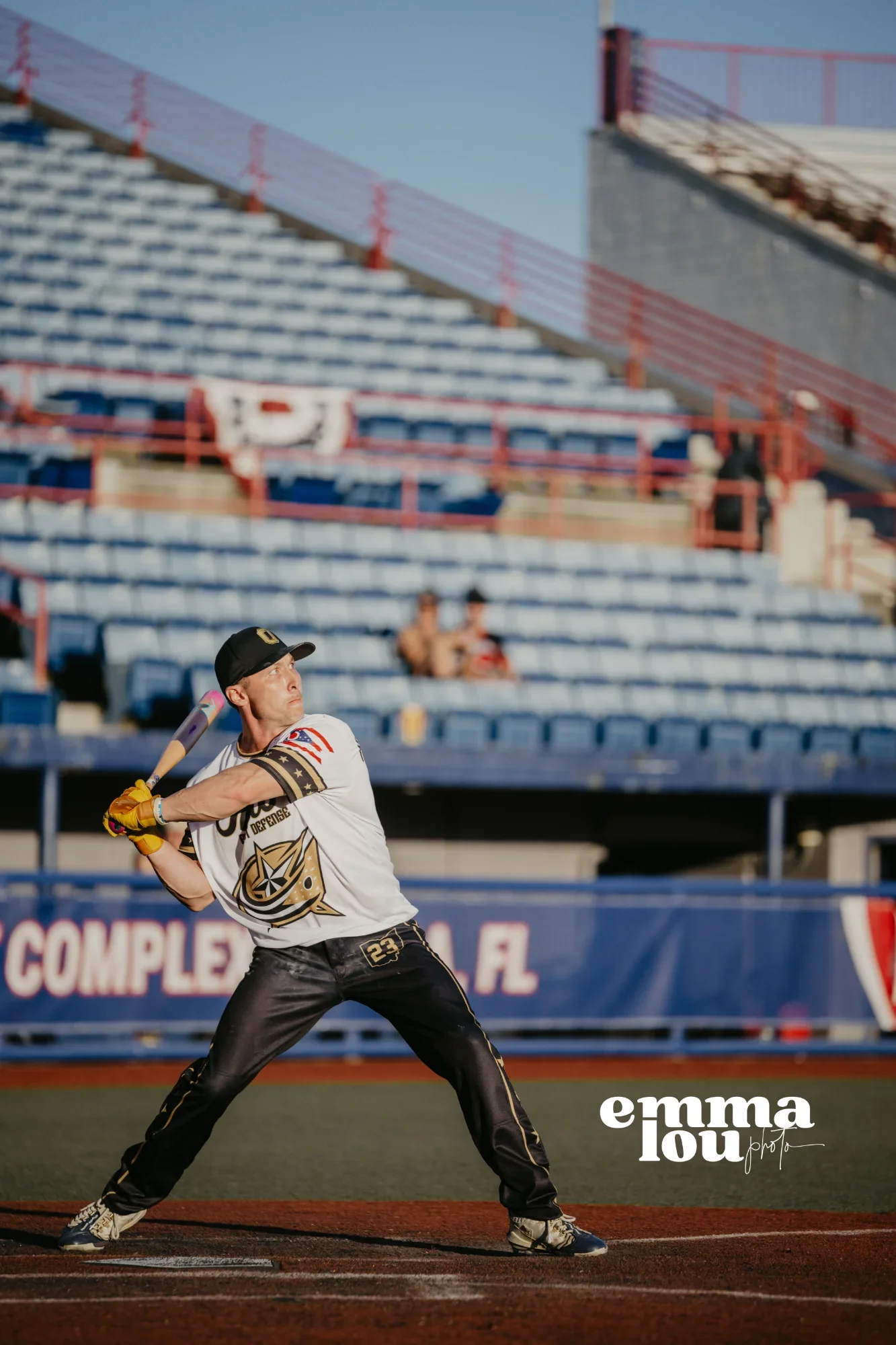 A baseball player in a black cap, white jersey, and black pants prepares to swing a bat on a baseball field with empty blue stadium seats in the background.