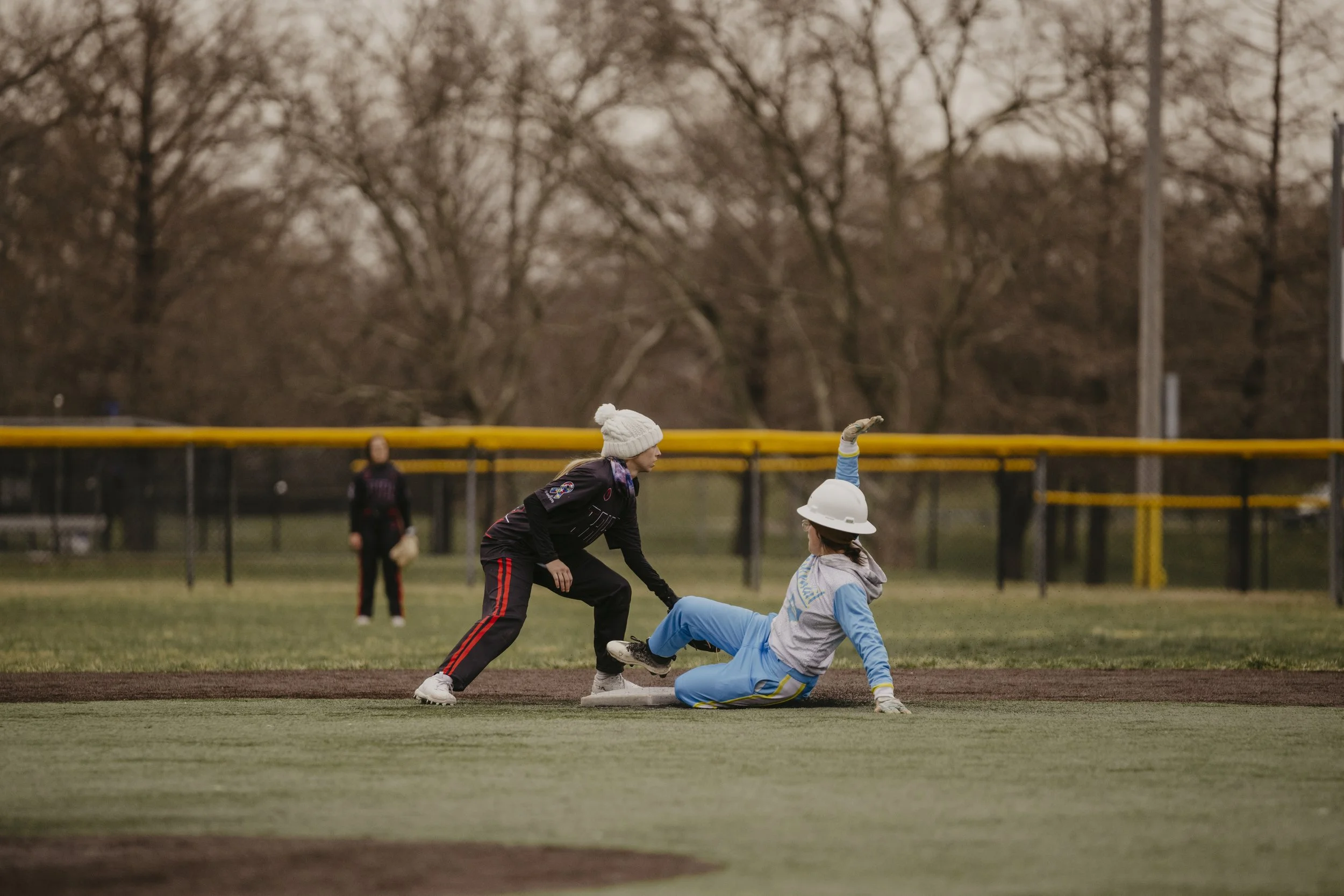 A woman sliding into a base during a softball game, with another woman in a black uniform attempting to tag her out, on a baseball field with a yellow fence and trees in the background.