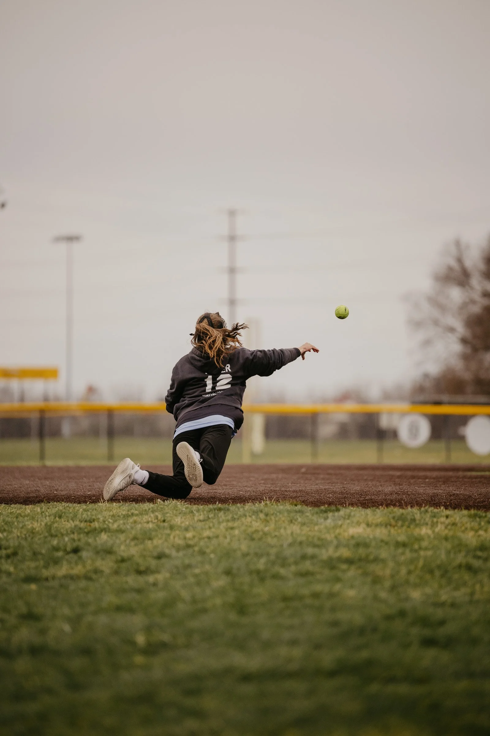 A softball player wearing a black sweatshirt and white cleats is diving on the field to catch a ball with one arm extended. The scene is at a baseball or softball field with a chain-link fence, green grass, and a cloudy sky.