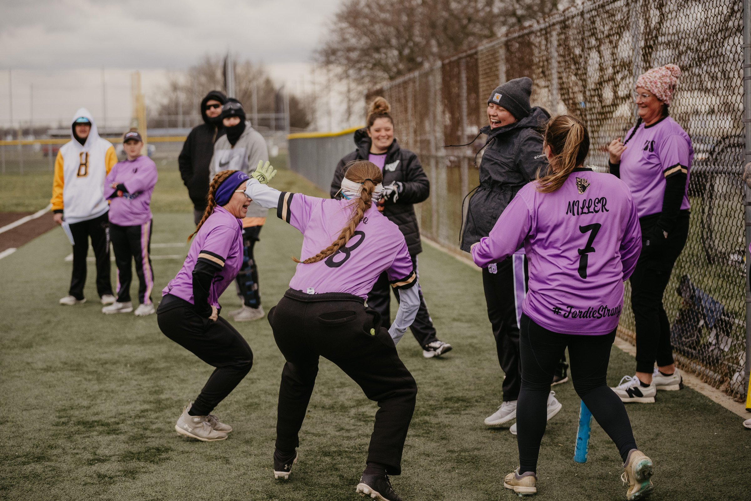A group of women and girls in purple softball uniforms on a field, celebrating and smiling, with some standing by a chain-link fence and others dancing or jumping.