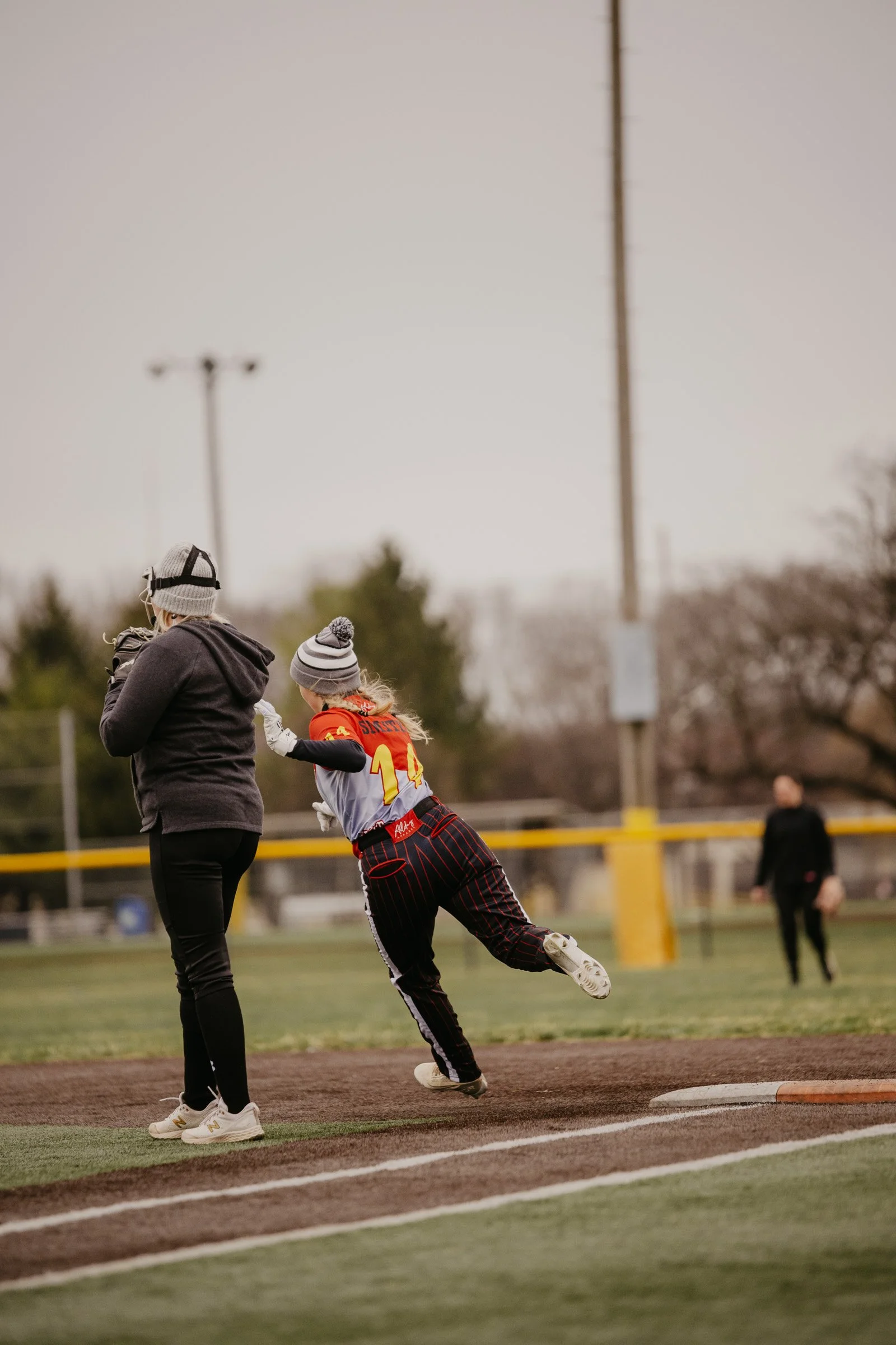 A young girl in a sports uniform running on a baseball field, with an adult woman nearby and another person in the background, during late afternoon or early evening.