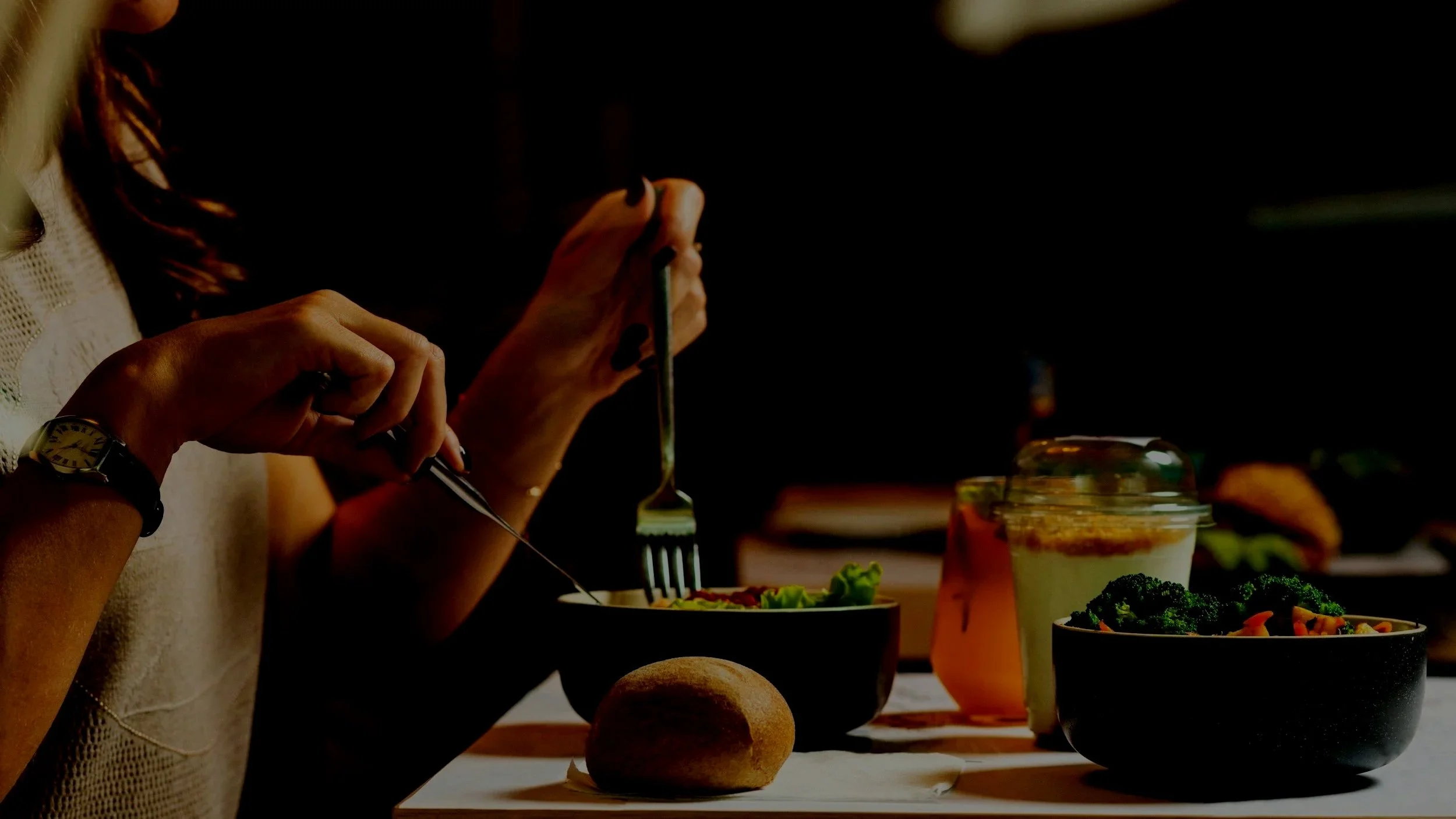 Person eating a salad with a fork at a table with bowls of salad, a bread roll, and drinks in a dimly lit setting.