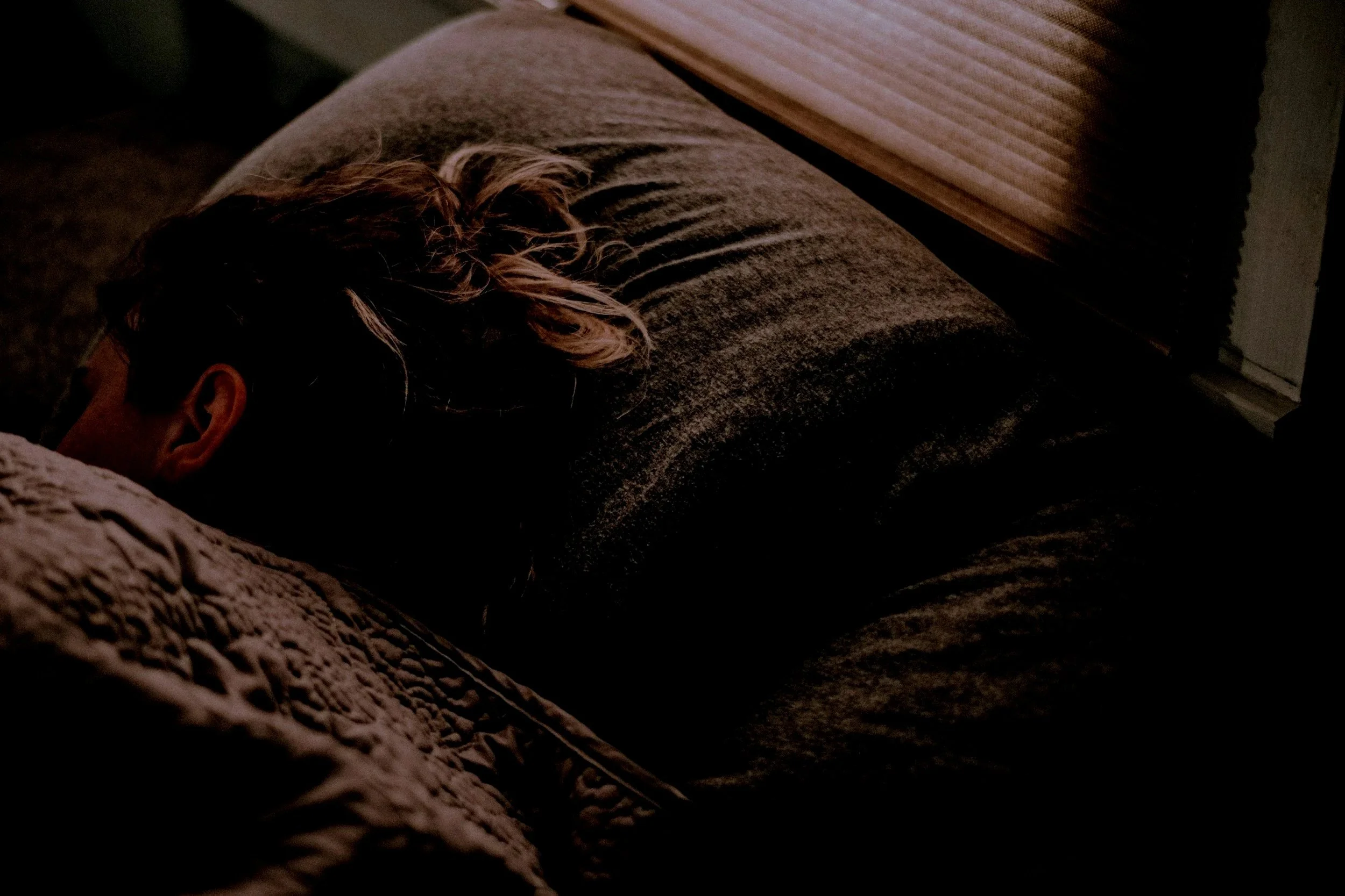 Person sleeping on a bed with dark bedding, turned to the side, with head resting on a pillow, in a dimly lit room.