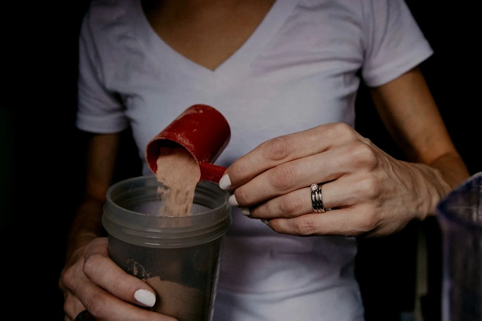 Woman in a white T-shirt pouring brown protein powder from a red scoop into a shaker bottle on the kitchen counter.