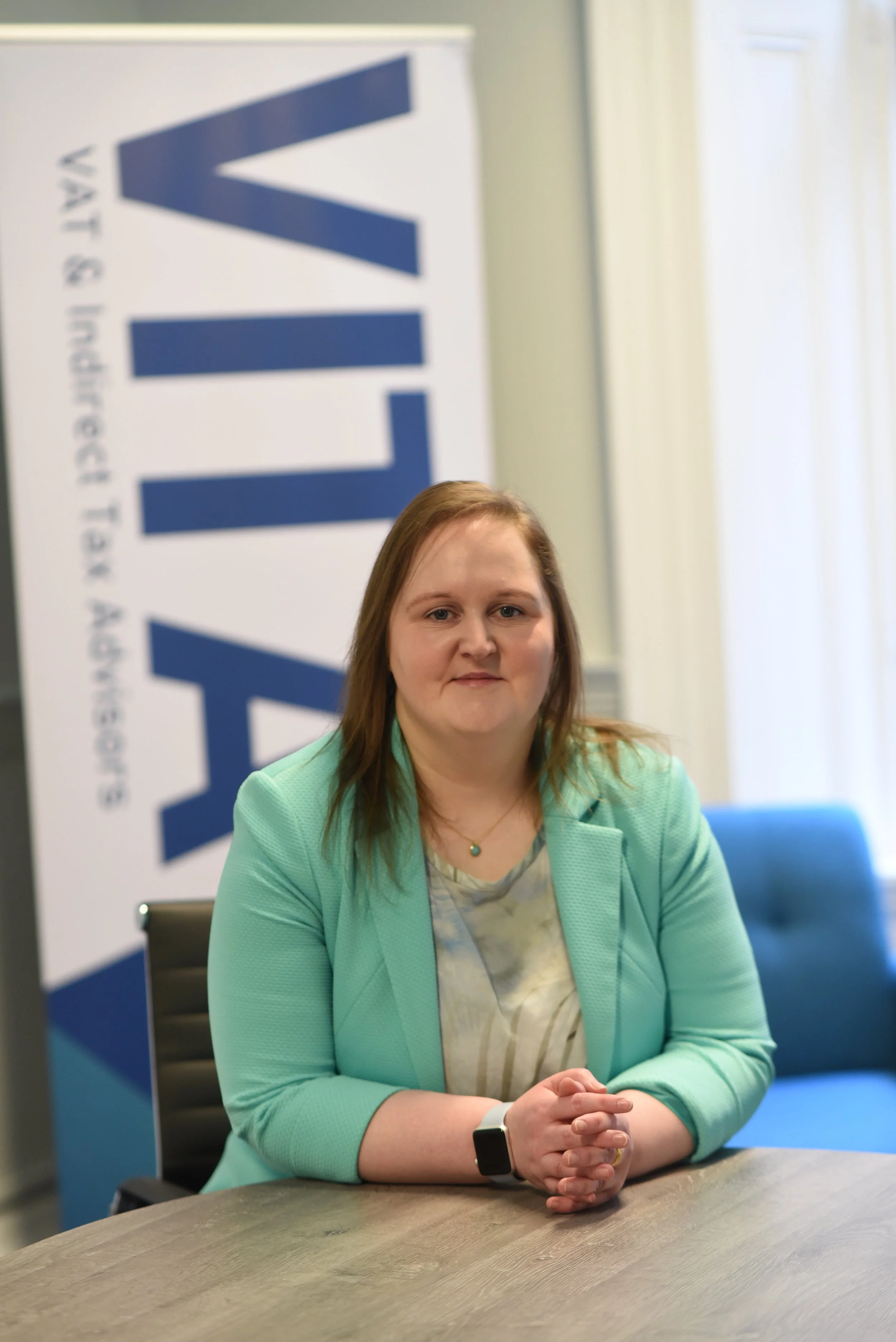 A woman with light skin and shoulder-length brown hair sitting at a table with hands clasped, wearing a mint green blazer and a smartwatch, in front of a blue and white sign that reads VOTE.