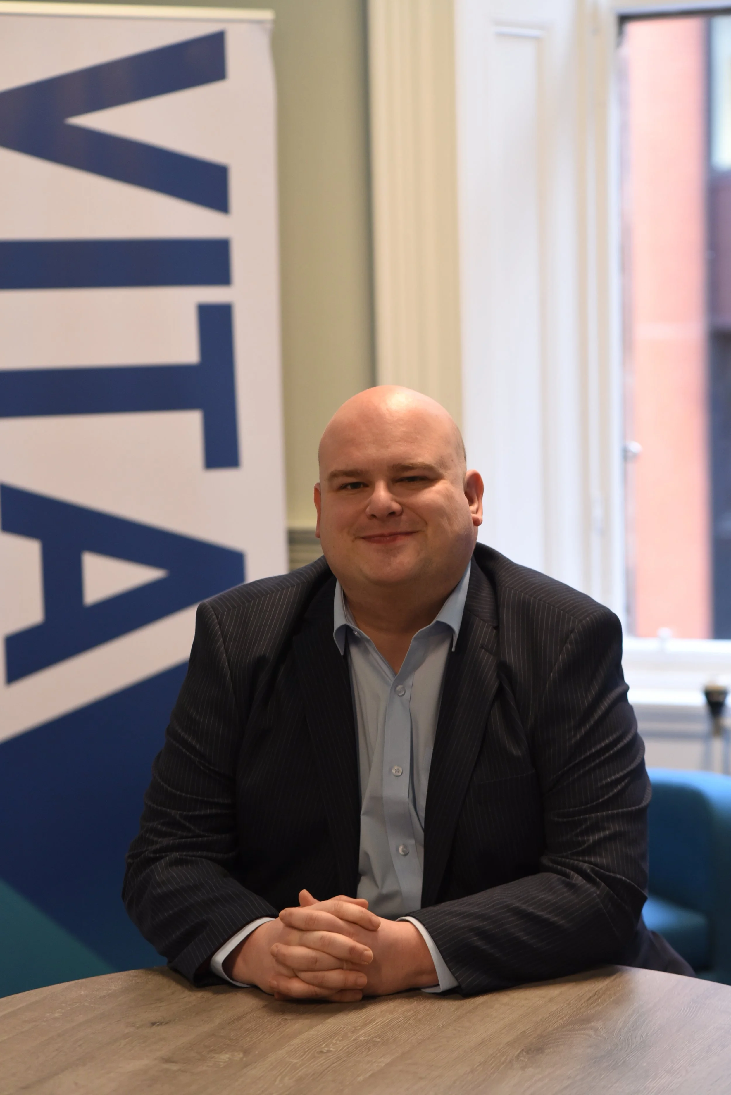 A man in a suit sitting at a table indoors with a large window behind him. There is a banner with the word 'WANT' partially visible on the left side of the image.