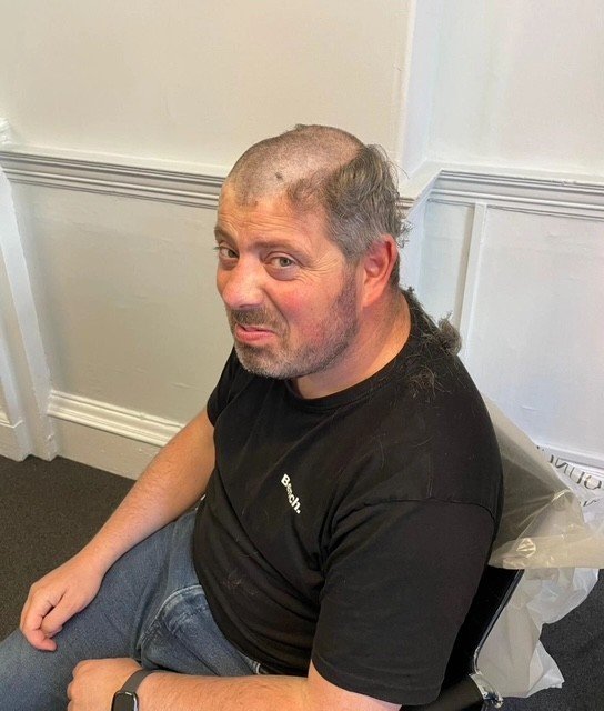 A man with a shaved head and facial hair sitting on a black chair outdoors, making a smirking facial expression.
