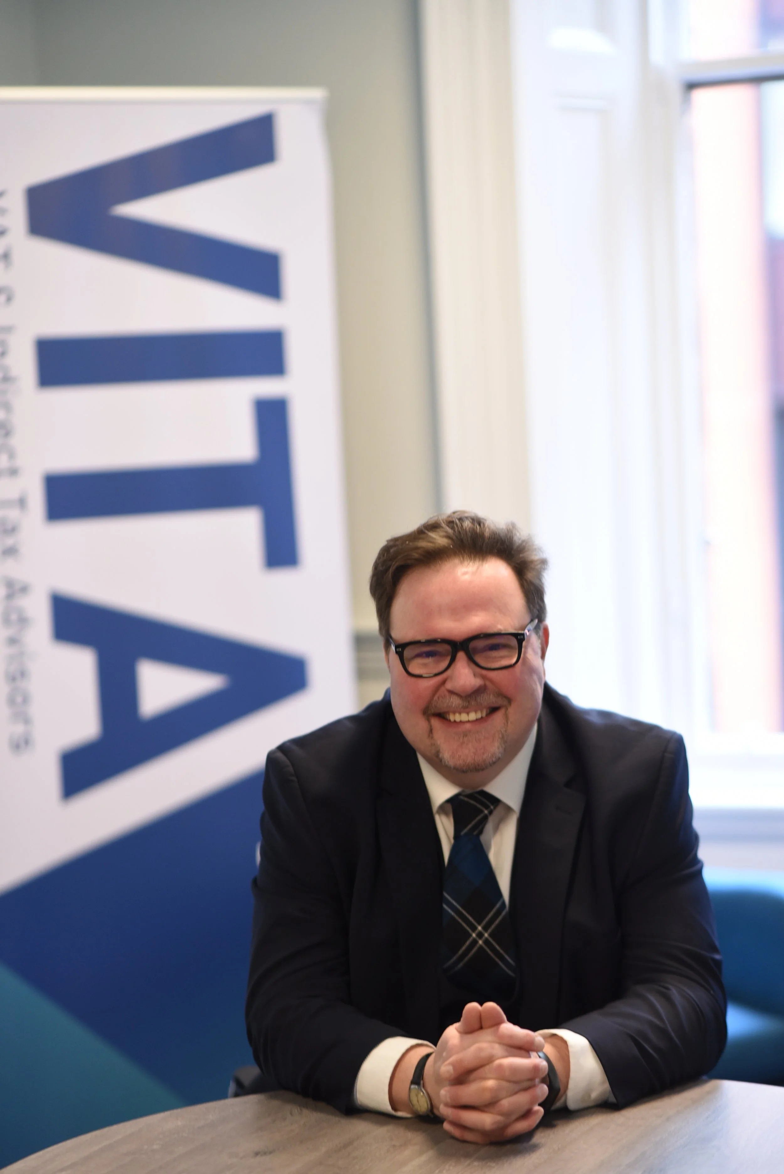 Man in a business suit, glasses, and a plaid tie smiling and sitting at a table in a room with a UNTY banner and a window in the background.