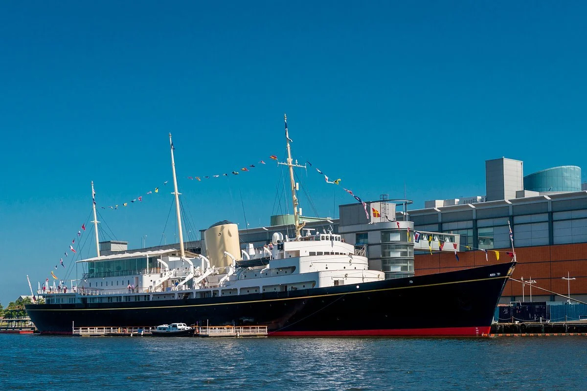 A large, vintage-style ship docked at a harbor with a modern building in the background. The ship has colorful flags strung along its masts and is reflected in the water.
