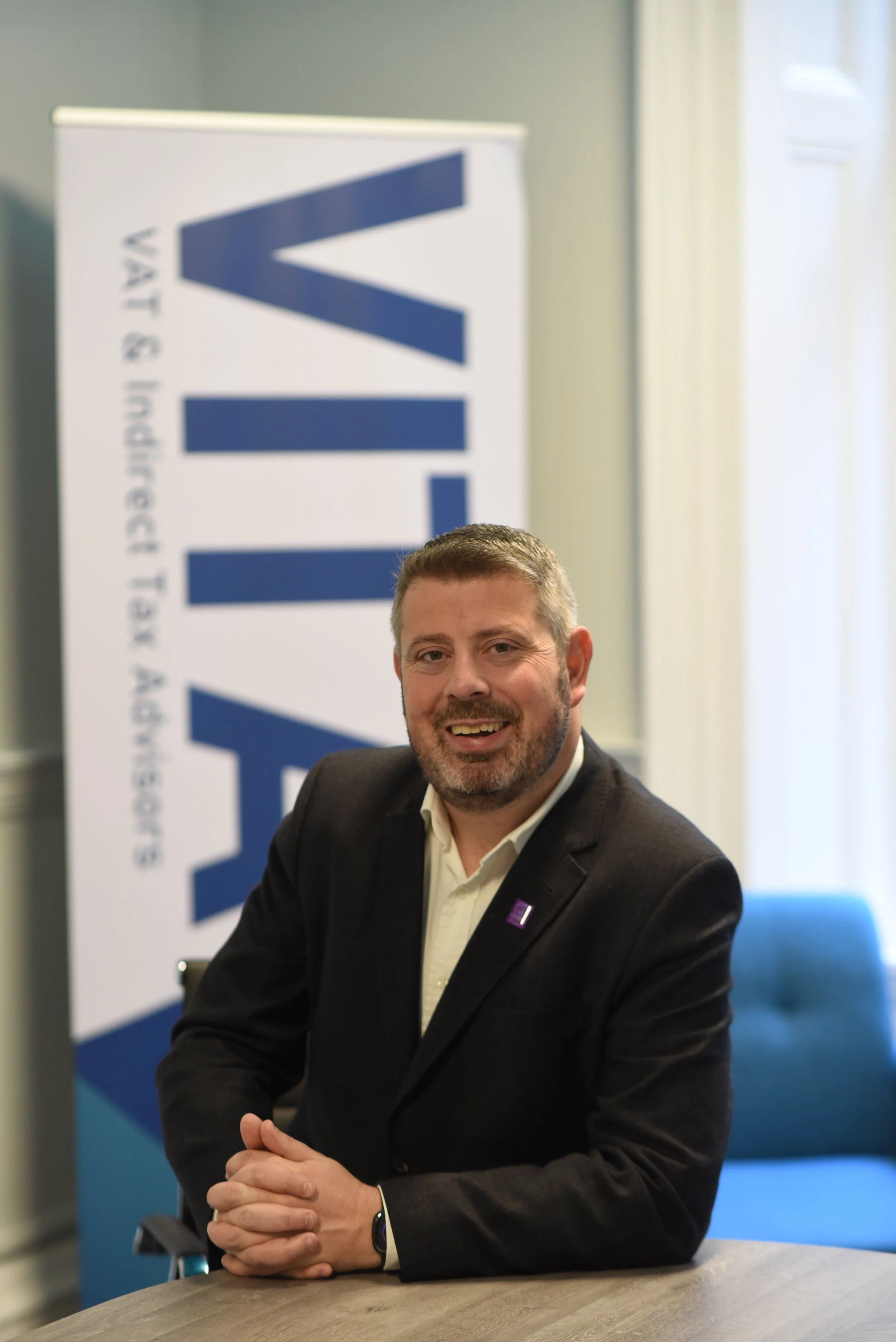 A man in a suit sitting at a table, smiling, with a banner in the background reading 'VITAL'.