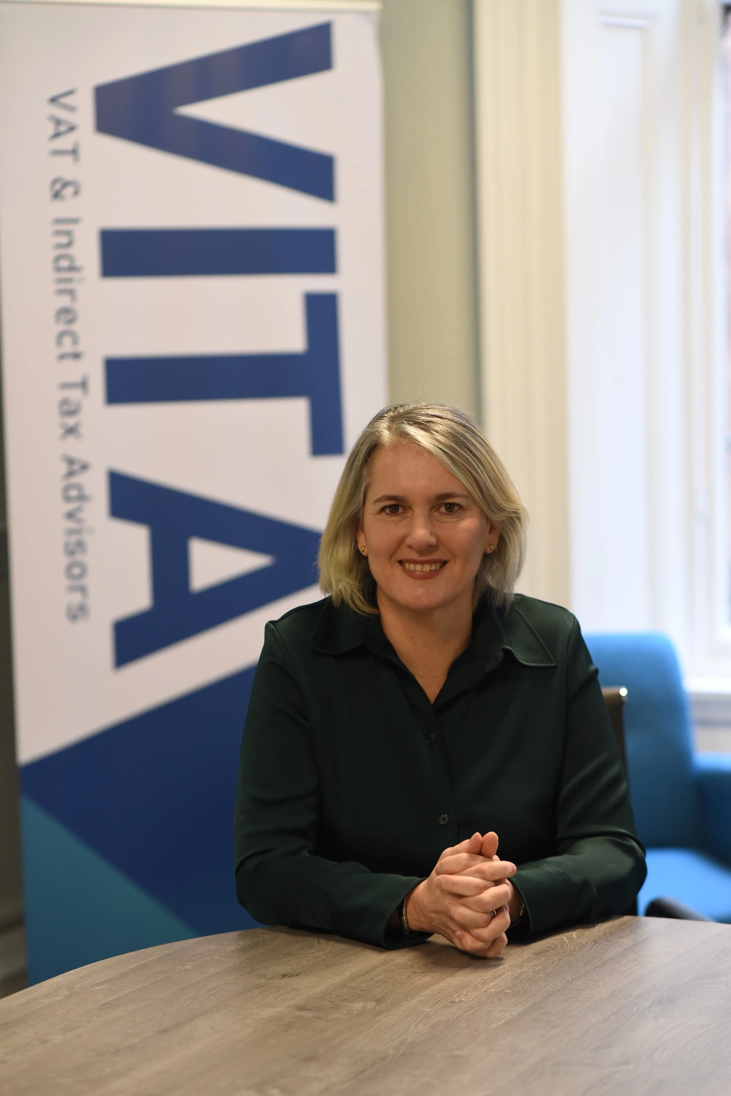 A woman with blonde hair and a black shirt sitting at a wooden table, smiling, with a blue and white banner in the background that reads 'VTV'.