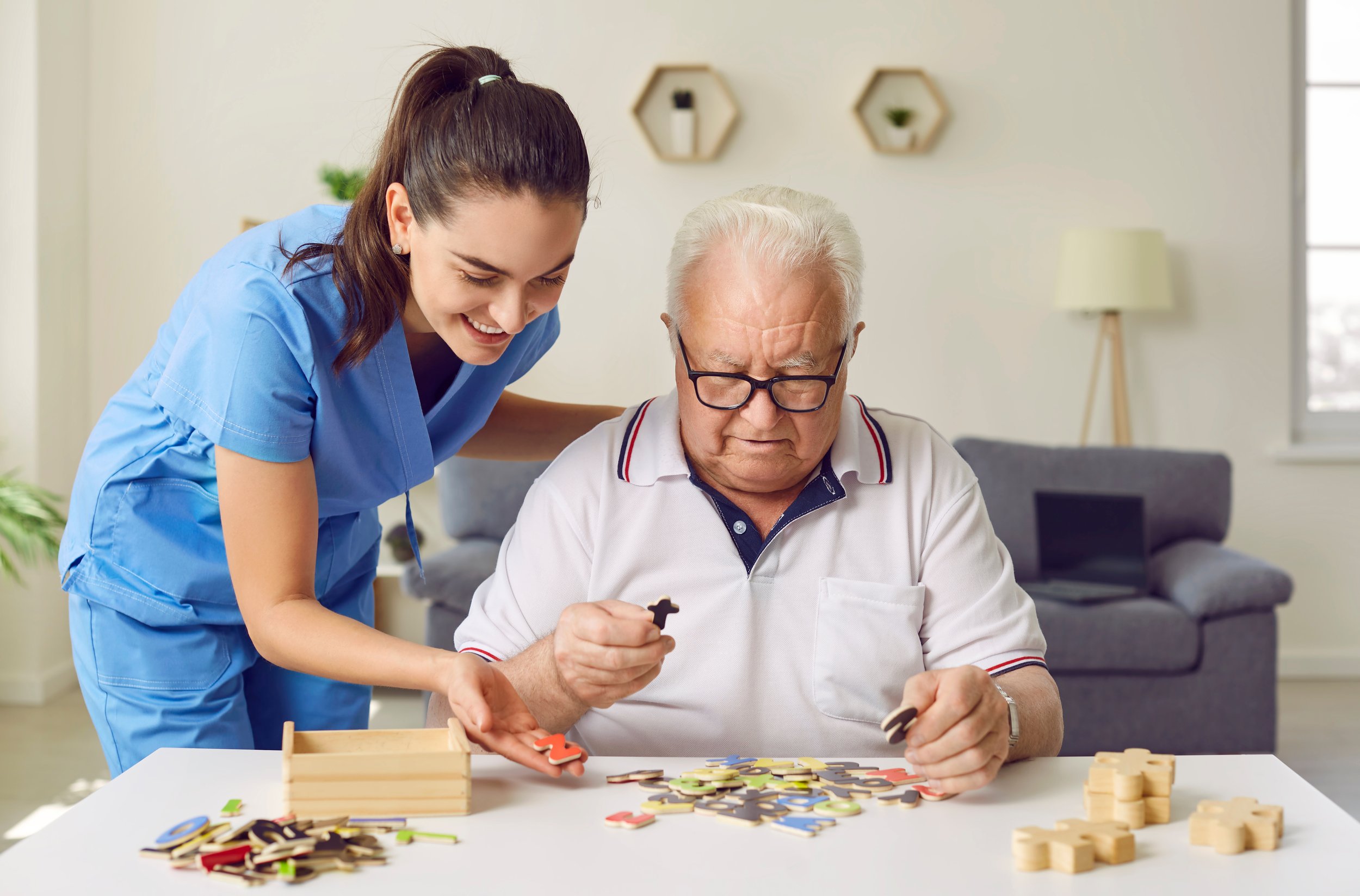 A young woman in medical scrubs smiles while playing a puzzle game with an elderly man in a white polo shirt and glasses at a table in a bright living room.