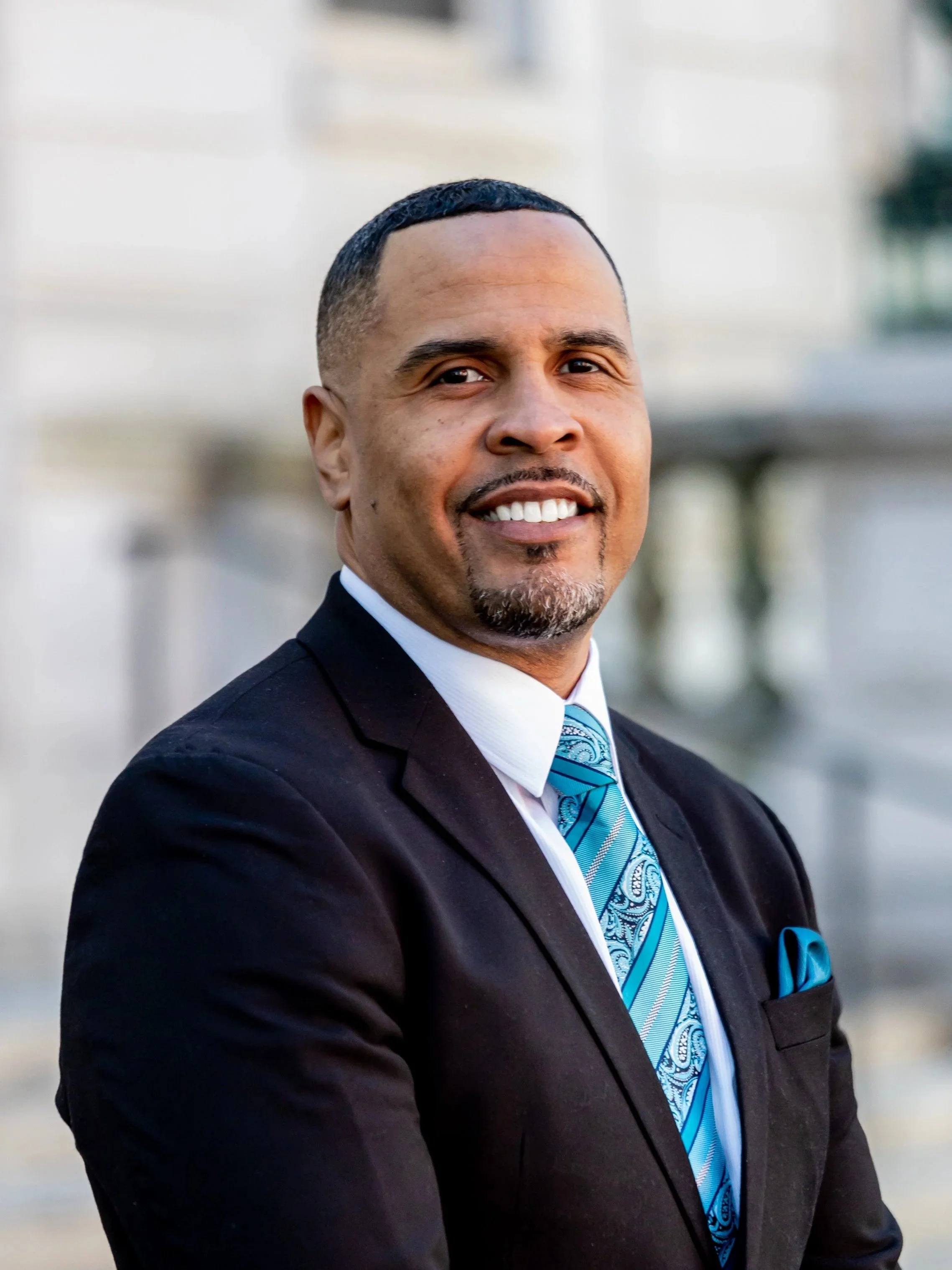 A man in a black suit, white shirt, and blue patterned tie, smiling outdoors with a blurred urban background.