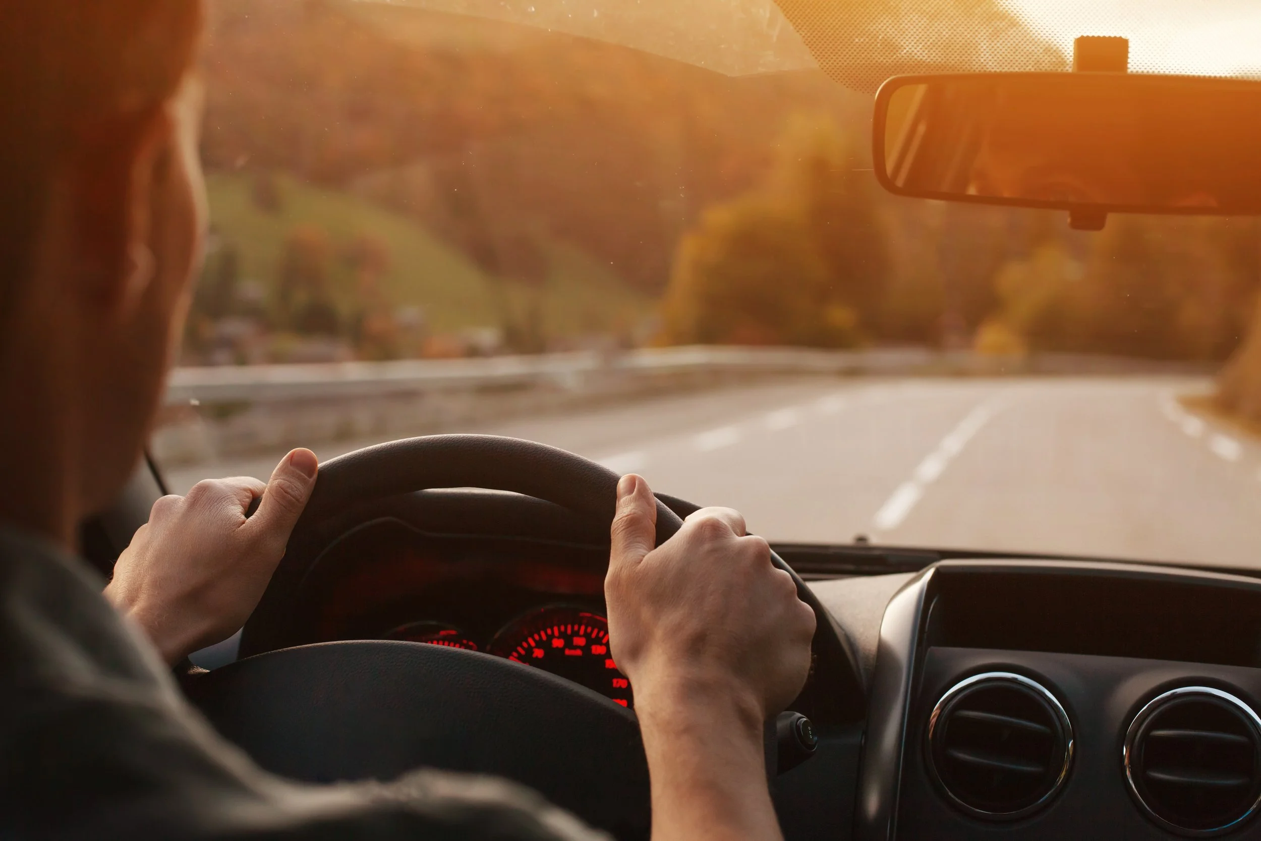 A person driving a car on a winding road during sunset with trees in the background.