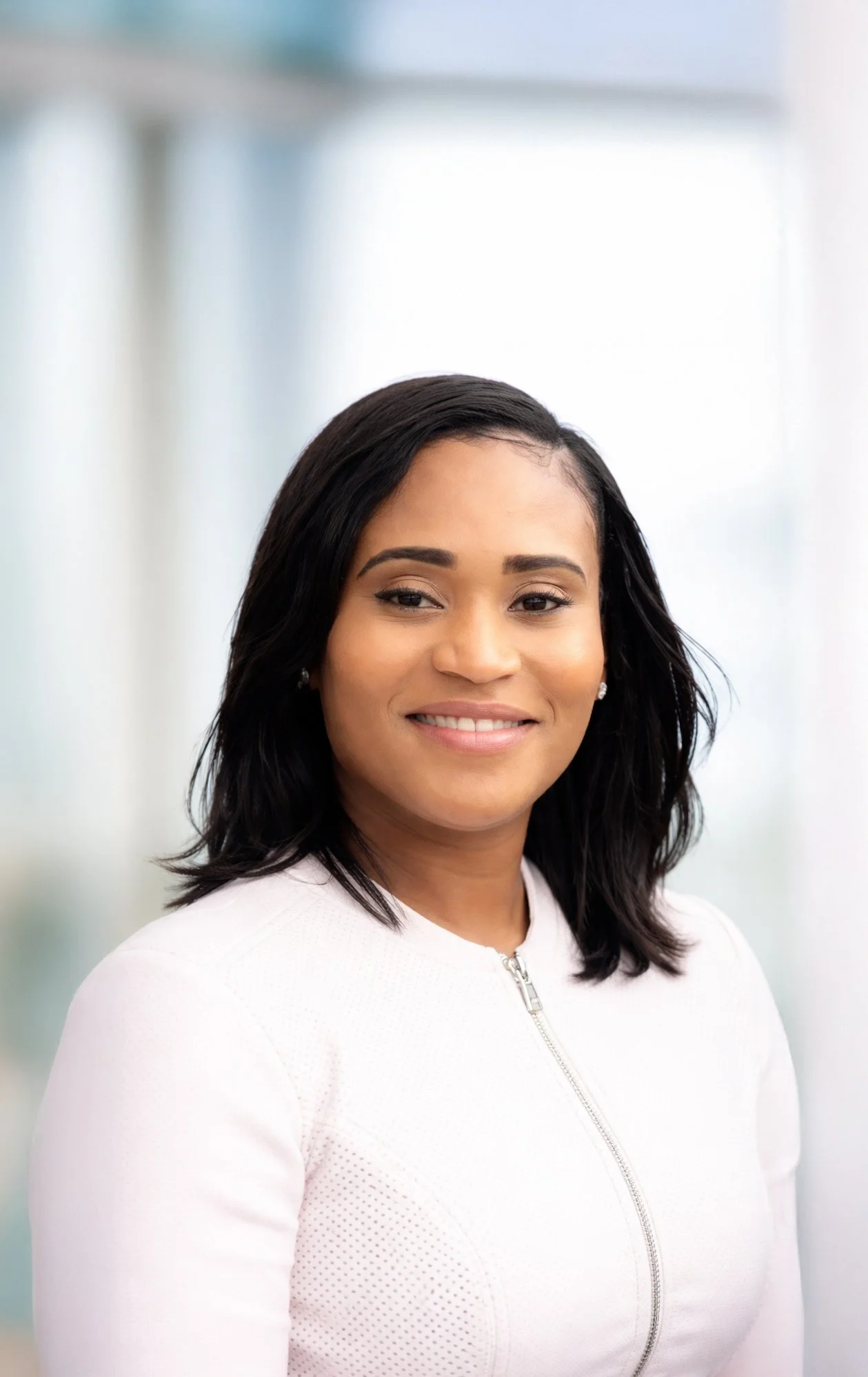 Portrait of a smiling woman with shoulder-length black hair, wearing a white zip-up top, standing indoors with a blurred background.