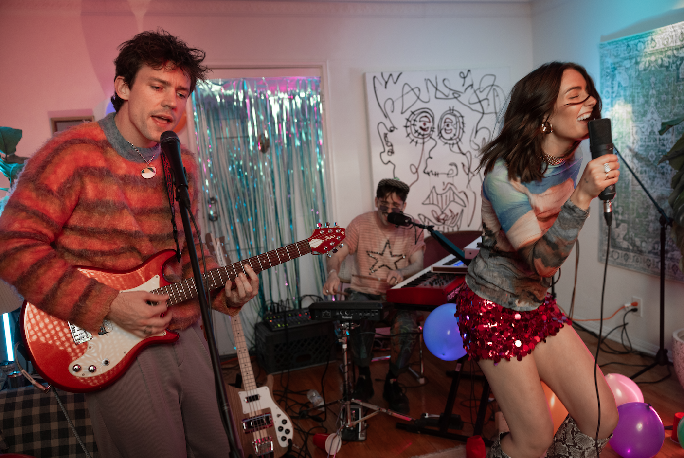 Three musicians performing at a lively indoor party, with colorful balloons and artwork in the background.