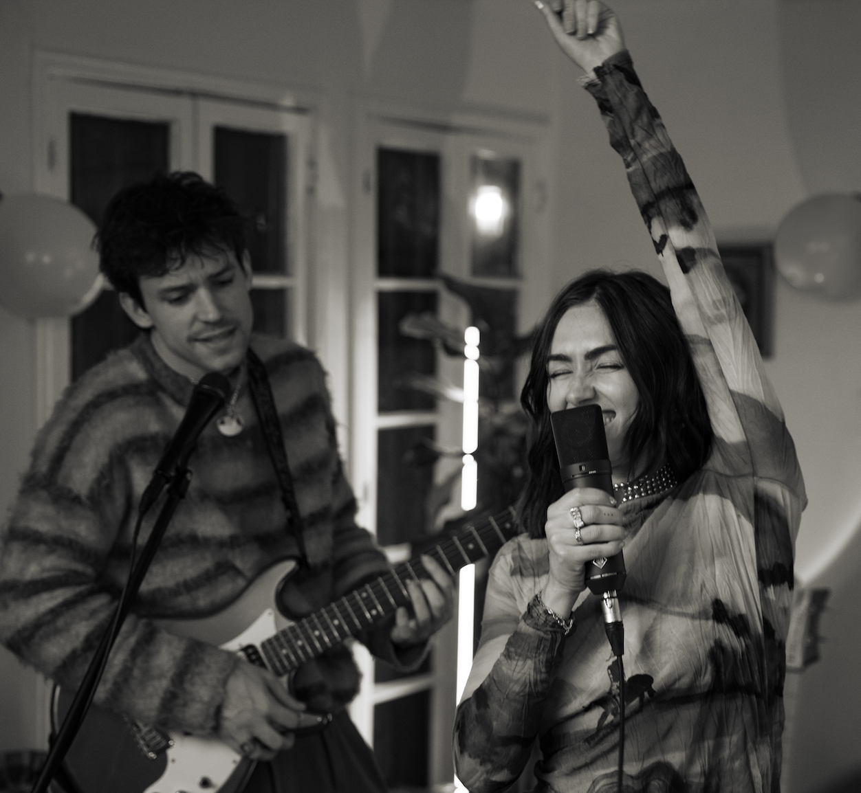 A woman is singing into a microphone with her eyes closed and arm raised while a man plays an electric guitar beside her, both performing indoors at a gathering.