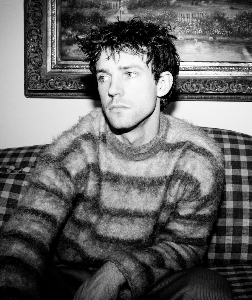 A young man with messy hair and light skin sitting on a checkered couch. He is wearing a fuzzy striped sweater and looking to his left, with a landscape painting in a decorative frame behind him.