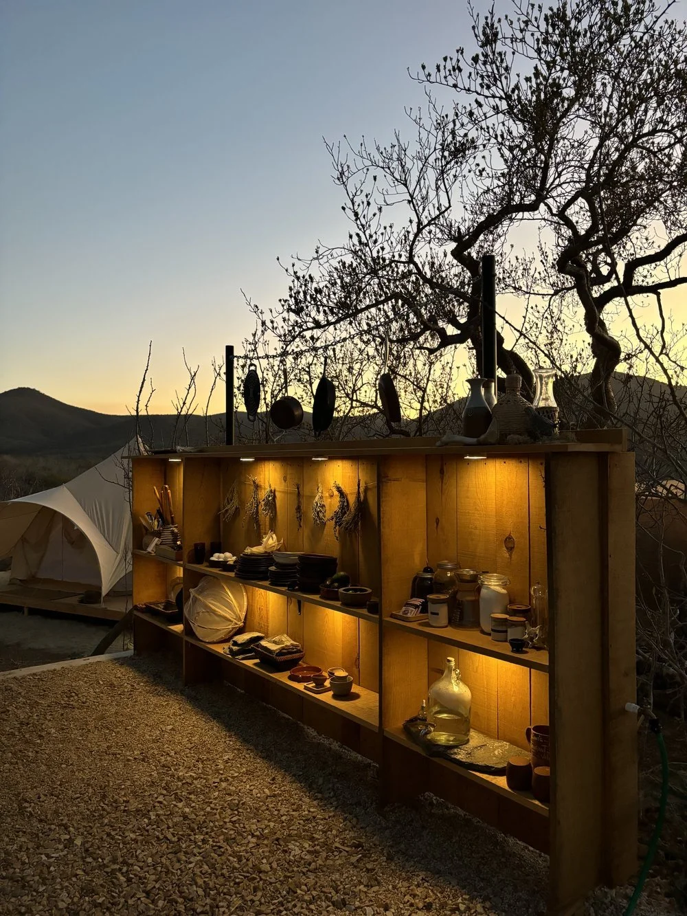 Outdoor display shelf with pottery and dried herbs, illuminated at dusk with a tent and mountain landscape in the background.
