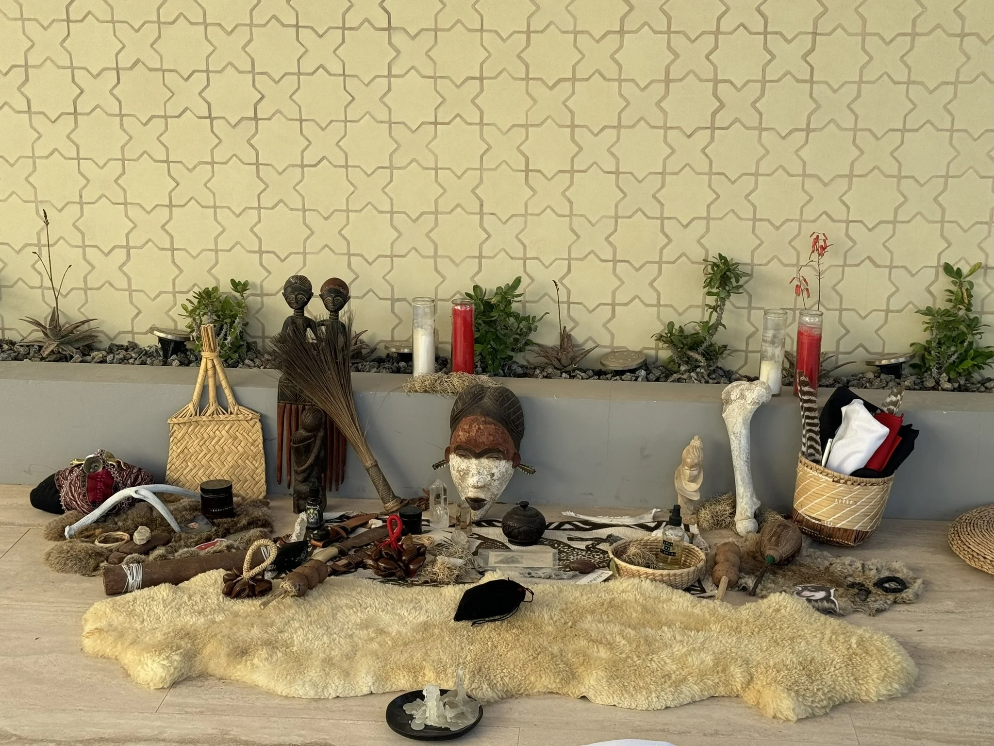 A collection of tribal and ritual artifacts arranged on a furry rug against a decorative wall. Items include carved wooden figures, animal bones, candles, woven baskets, masks, and various natural and crafted objects.