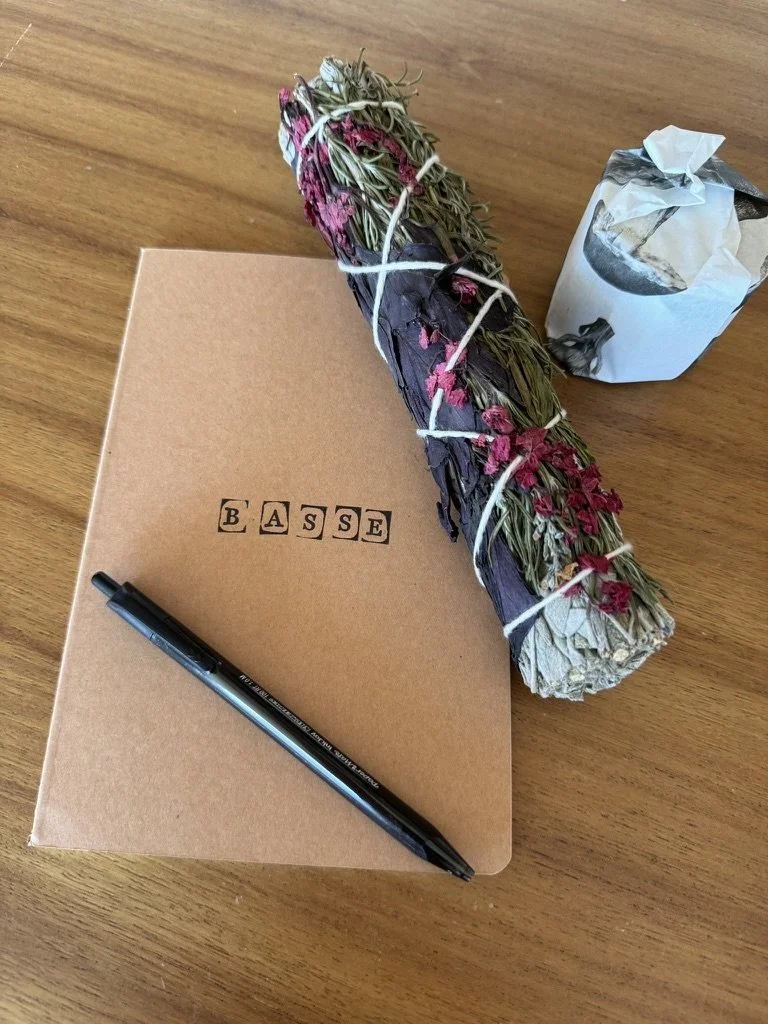 A bundle of dried herbs or flowers tied with white string, a closed toilet paper roll, a closed brown cardboard box labeled 'BASE' with black letter stamps, and a black pen on top of the box placed on a wooden surface.