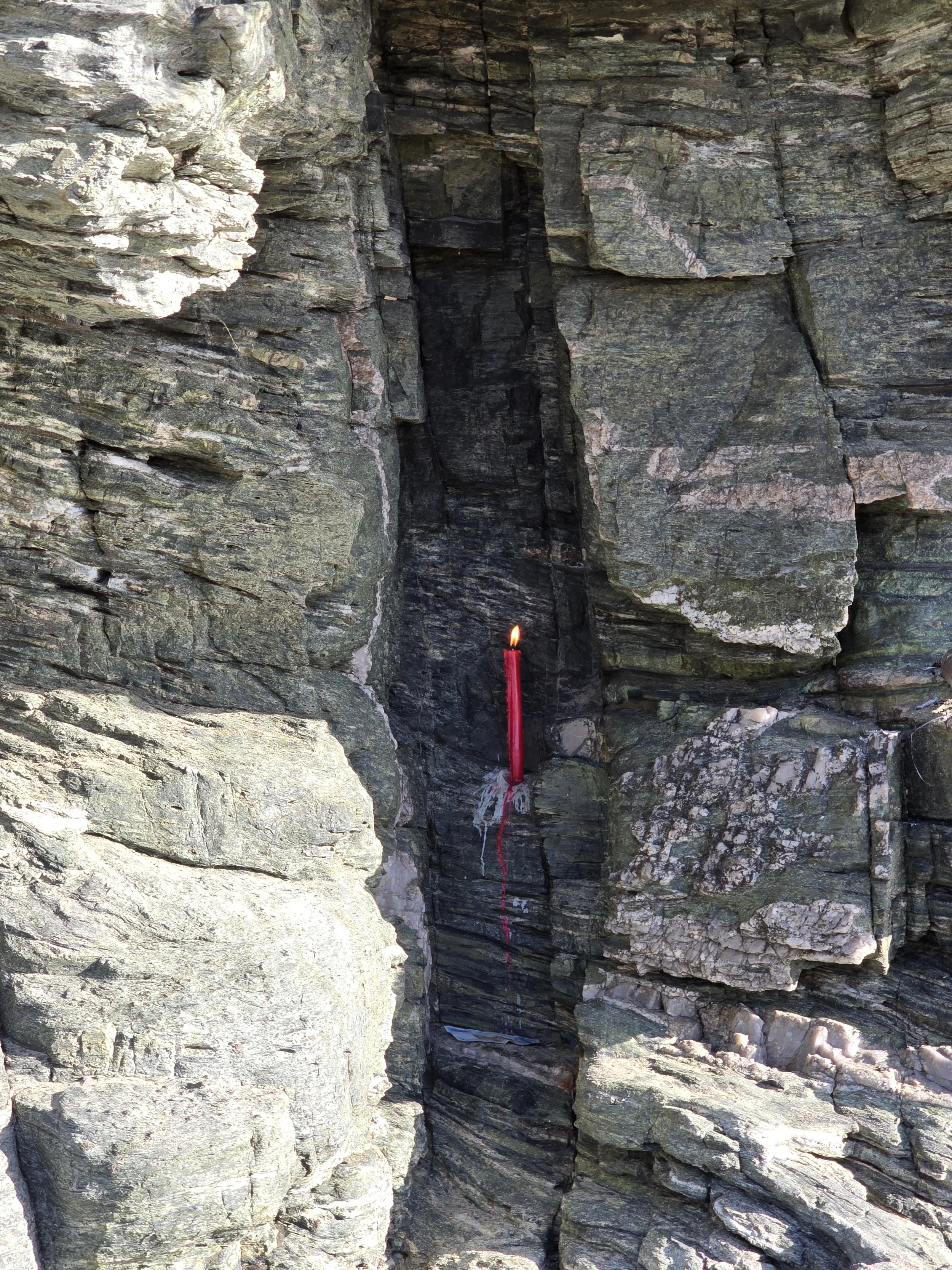 A lit red candle in a small crevice between large, layered rock formations with some wax dripping down.