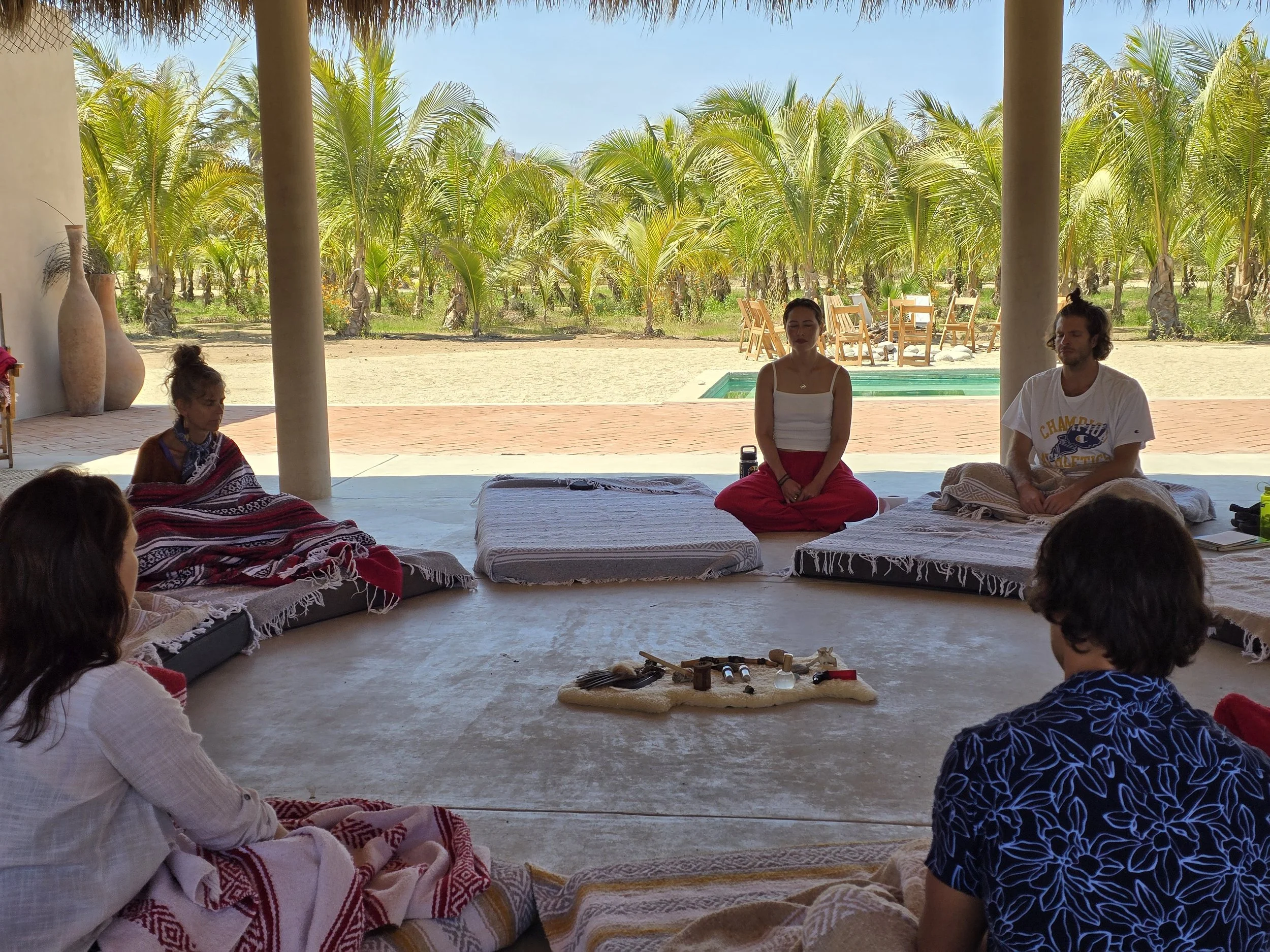 A group of people engaged in a meditation session indoors with an open view of palm trees and a swimming pool outside.