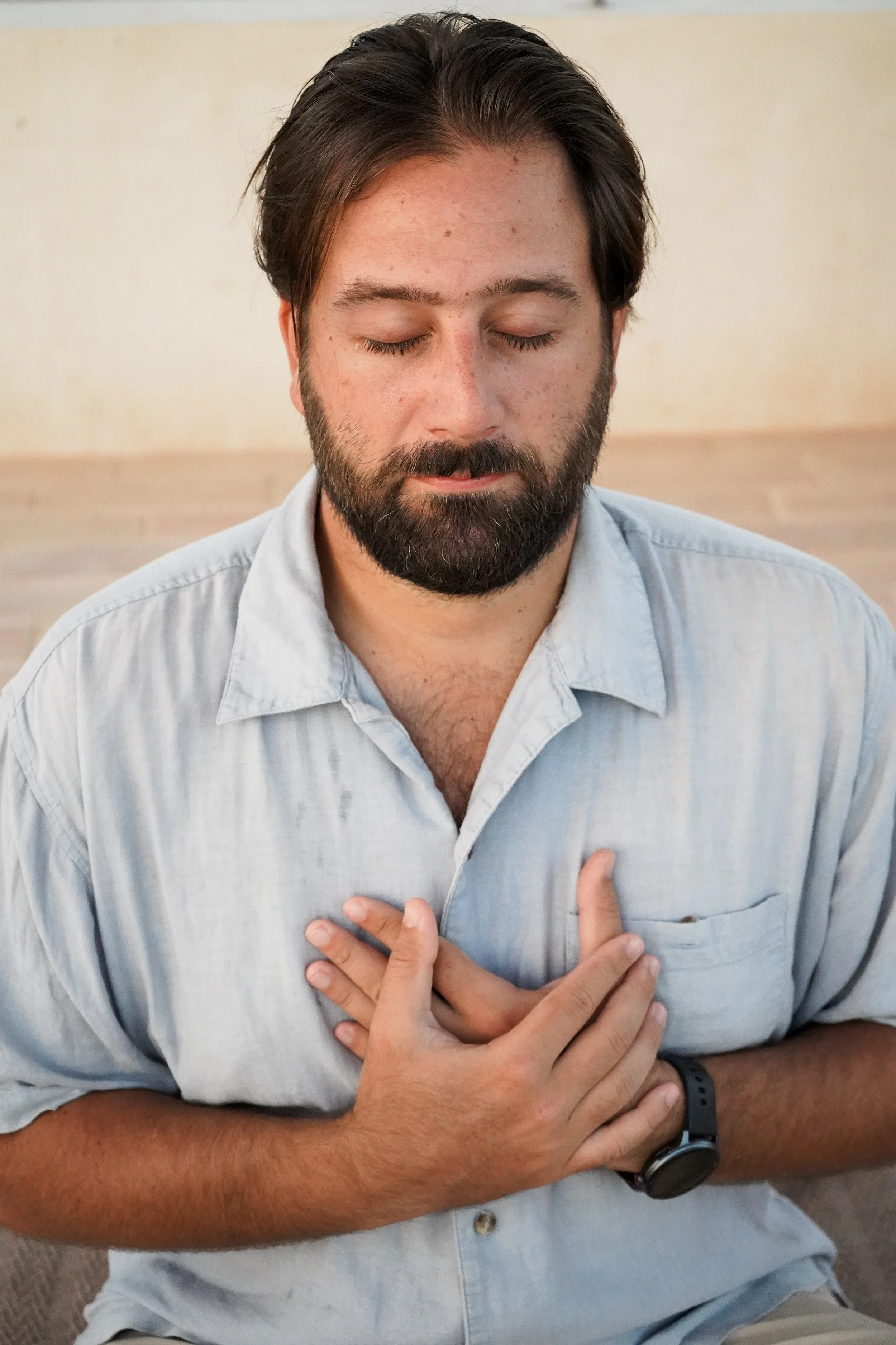 A man with brown hair, a beard, and closed eyes, holding his chest with one hand and appearing to experience chest discomfort.