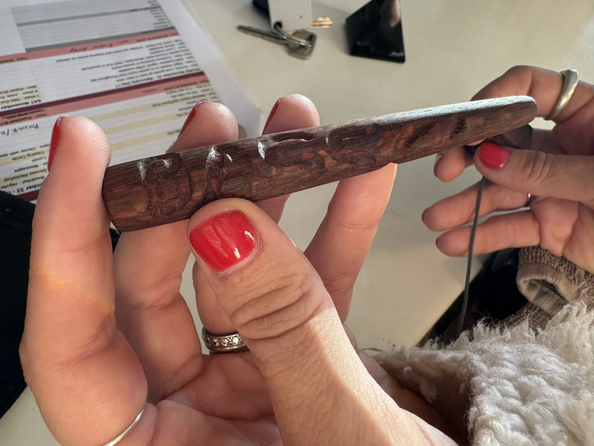 Close-up of hands holding a carved wooden object and using a crochet hook, with a background including a document and a small mirror.