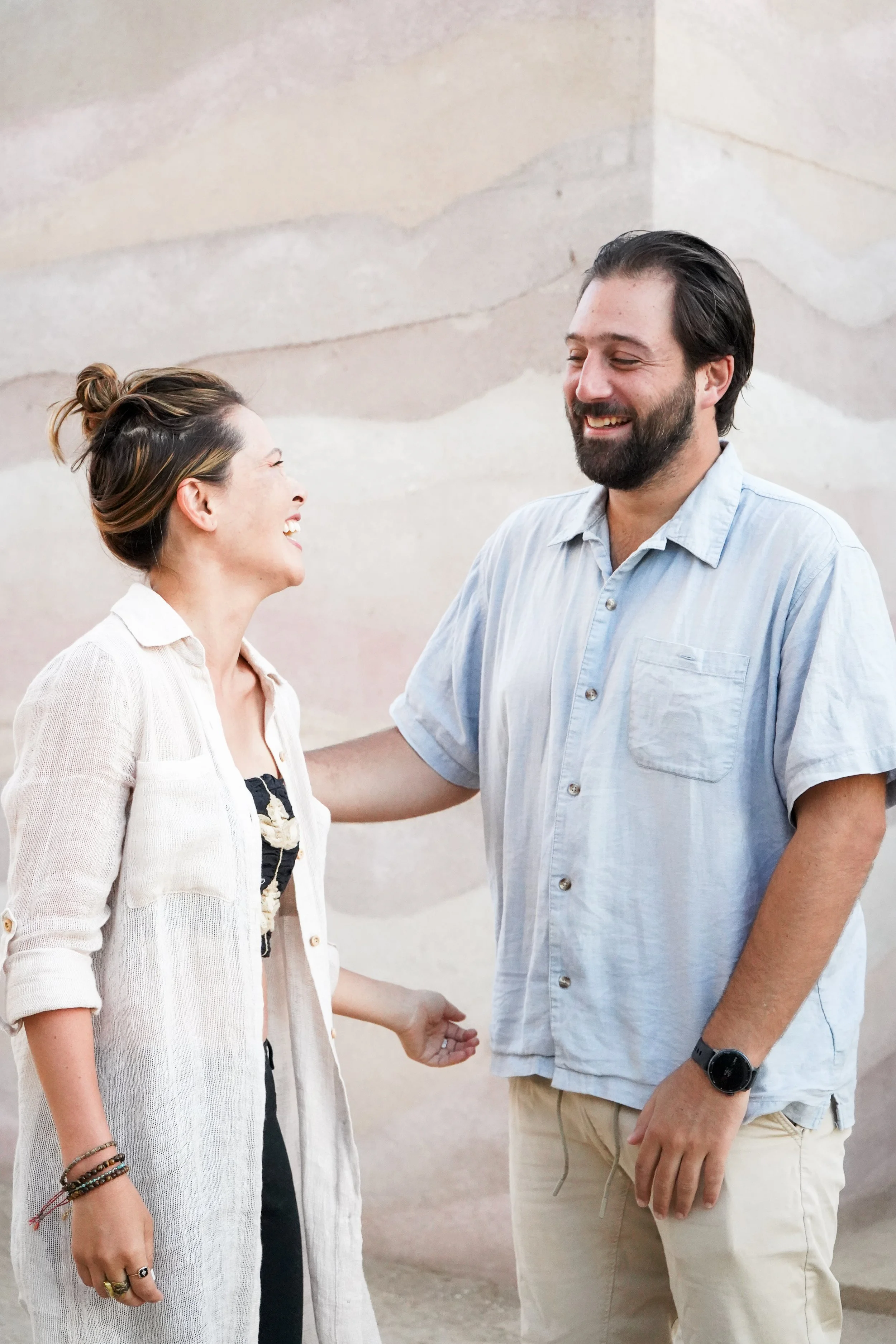 A man and woman smiling and laughing, standing close together against a textured wall. The woman has her hair in a bun, is wearing a light-colored cardigan, and has bracelets and rings. The man has a beard, is wearing a light blue short-sleeve shirt, beige pants, and a black watch.