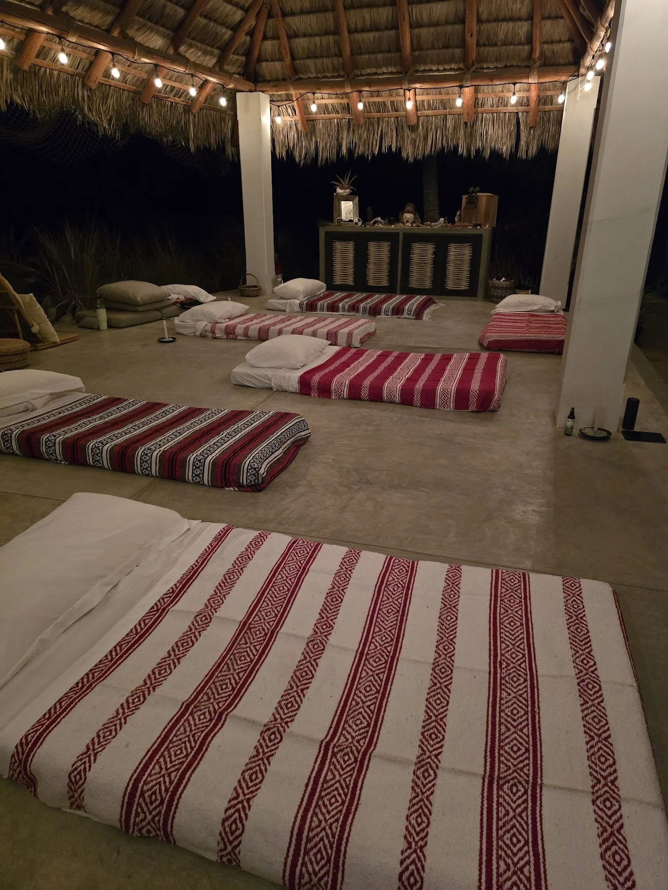 Outdoor open-air space with a thatched roof, illuminated by string lights, featuring several traditional patterned mats laid out on the floor with pillows at one end, and a wooden cabinet against the back wall.