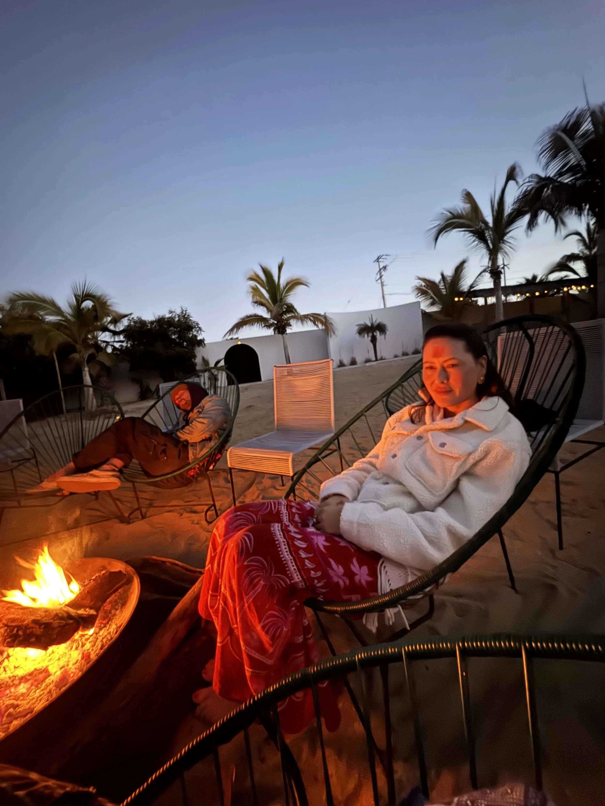 Two women sitting on outdoor chairs near a campfire on a sandy area, with palm trees and a dusk sky in the background.