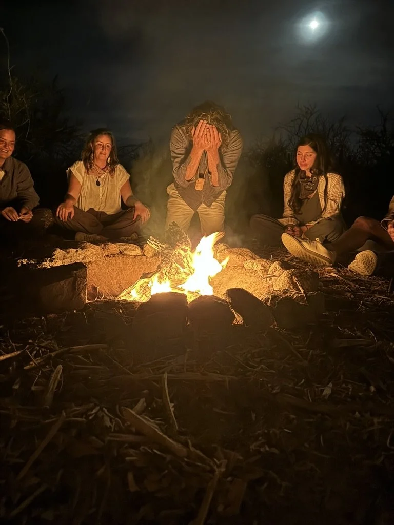A group of people sitting outside at night around a campfire, with one person in the center covering their face, illuminated by the flames under a cloudy moonlit sky.
