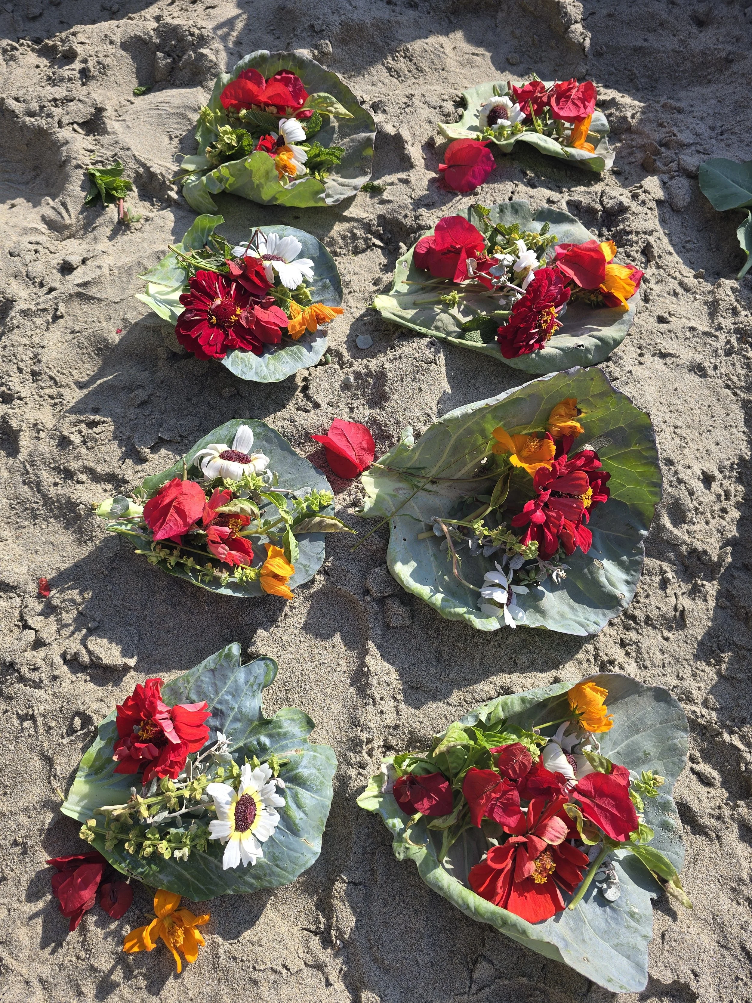 Assorted flower arrangements placed on sandy ground, featuring red, white, yellow, and orange flowers on large green leaves.
