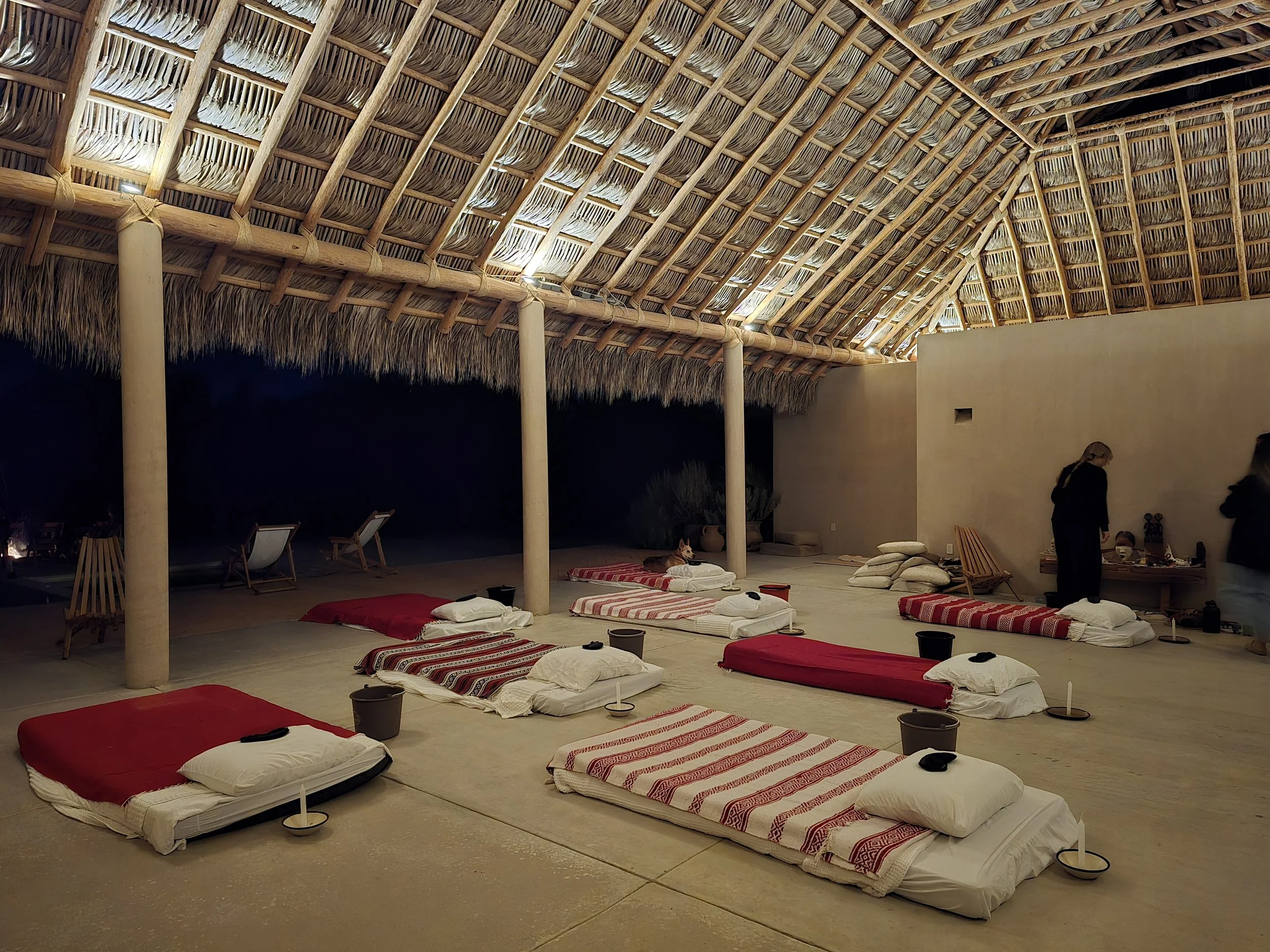 An outdoor open-air space with traditional mattresses laid on the floor, each topped with white pillows and red or striped blankets, under a thatched roof with wooden support beams. There are candles and small buckets beside each bed, and a seated dog near the back. Two women are standing near a wall with various items on a low table.