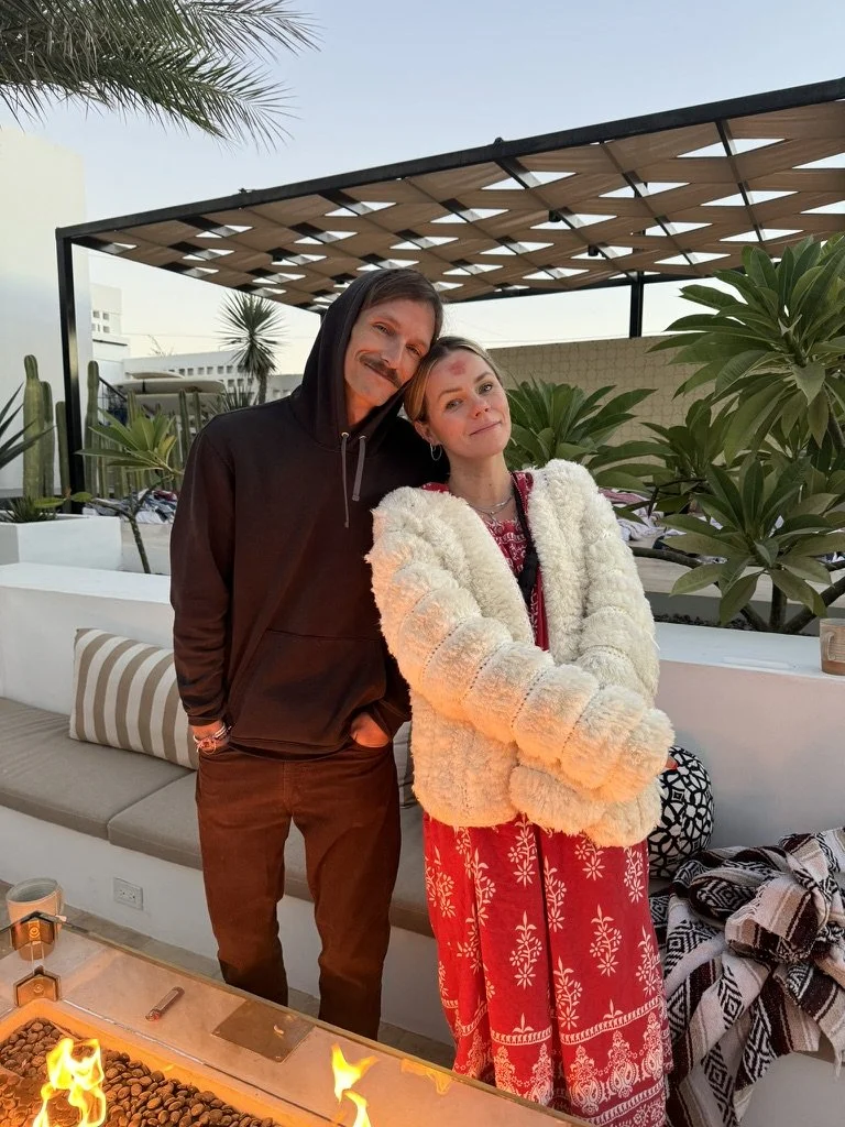 A man and woman are standing close together outdoors during sunset, smiling. The man is wearing a black hoodie and brown pants. The woman is wearing a fluffy white jacket and a red patterned dress. There is tropical vegetation and a modern outdoor structure in the background, and a fire pit or tabletop fire feature in the foreground.