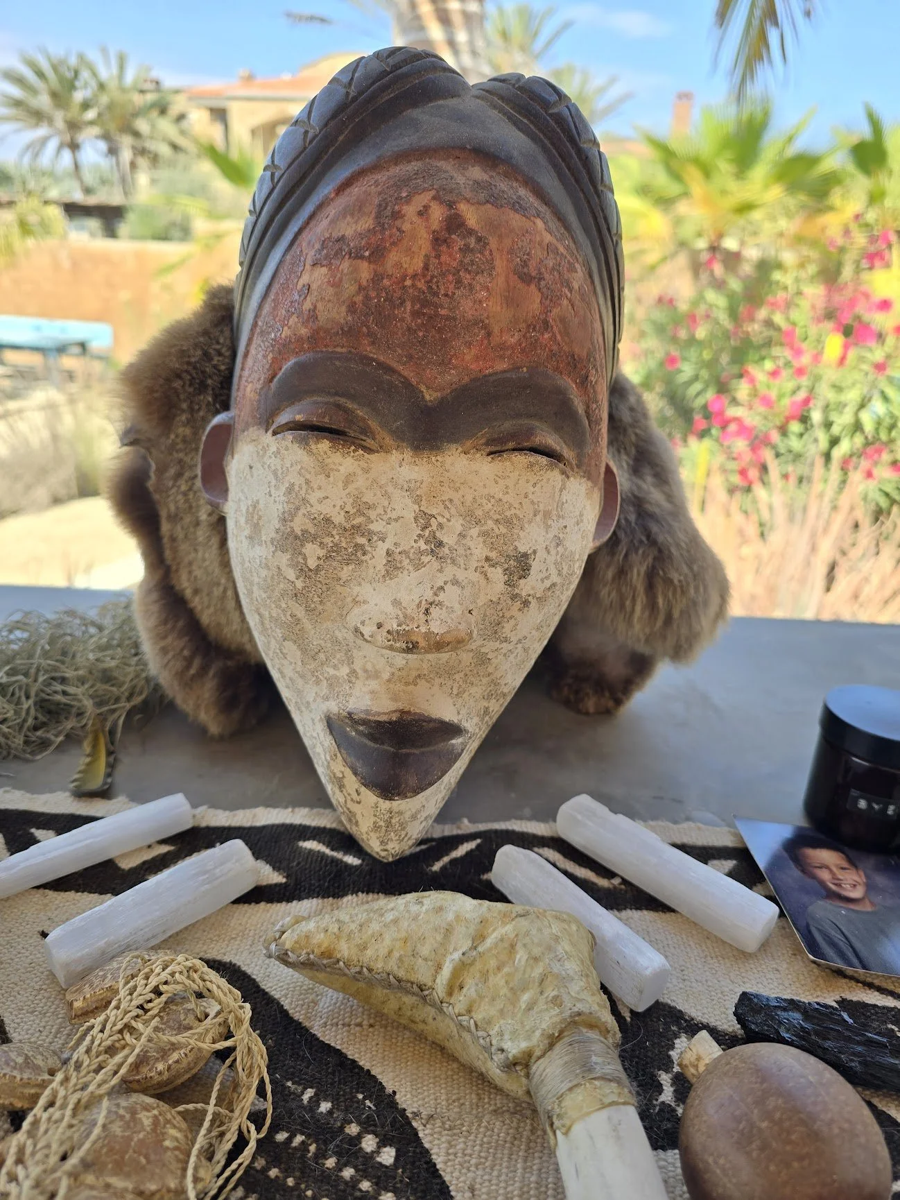 An African tribal mask with a weathered face, dark lips, and closed eyes, surrounded by various objects including white bones, a horn, and stones, outdoors with trees and plants in the background.